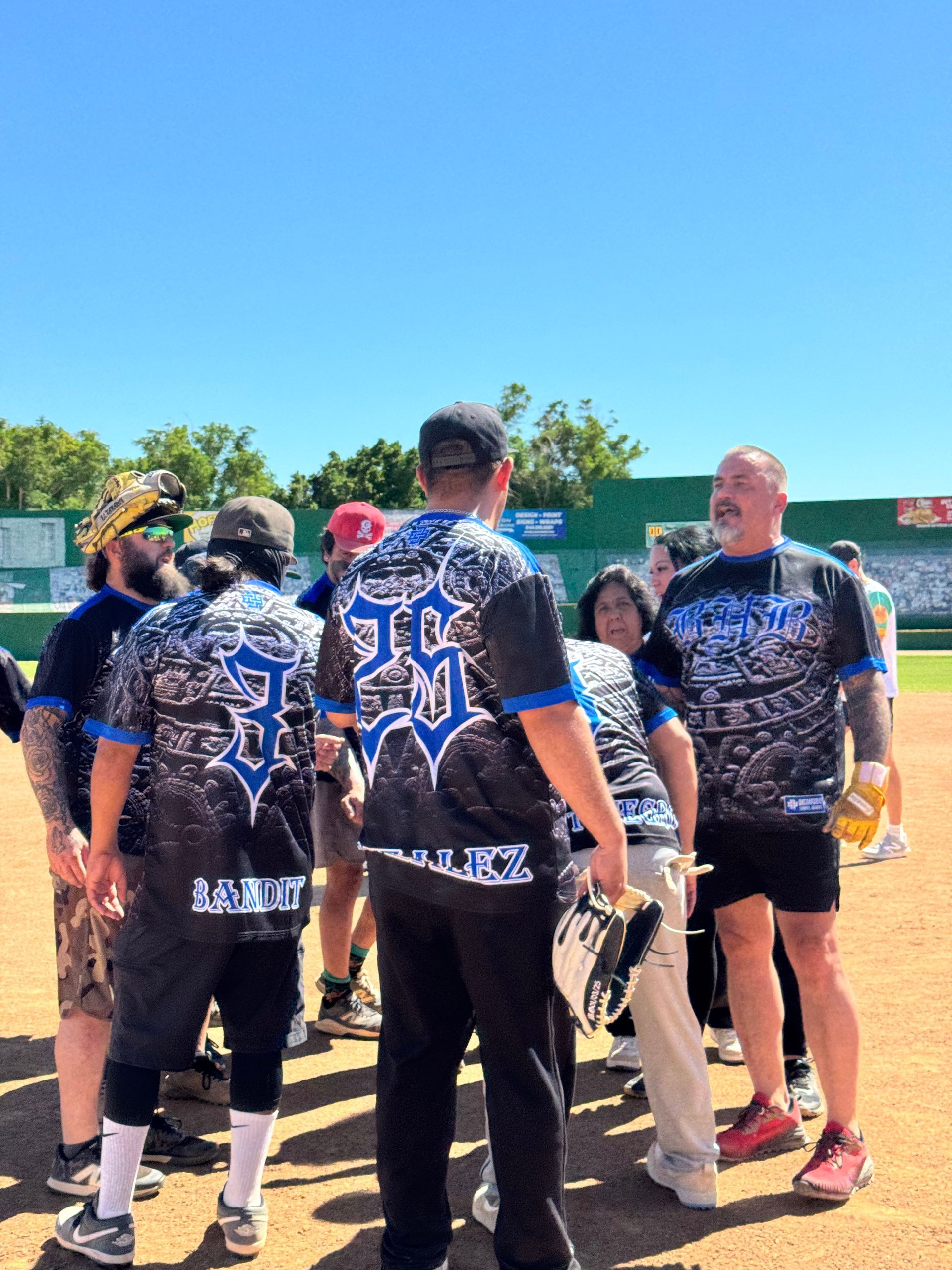 Softball players in blue camo jerseys huddle on a field under a clear blue sky.