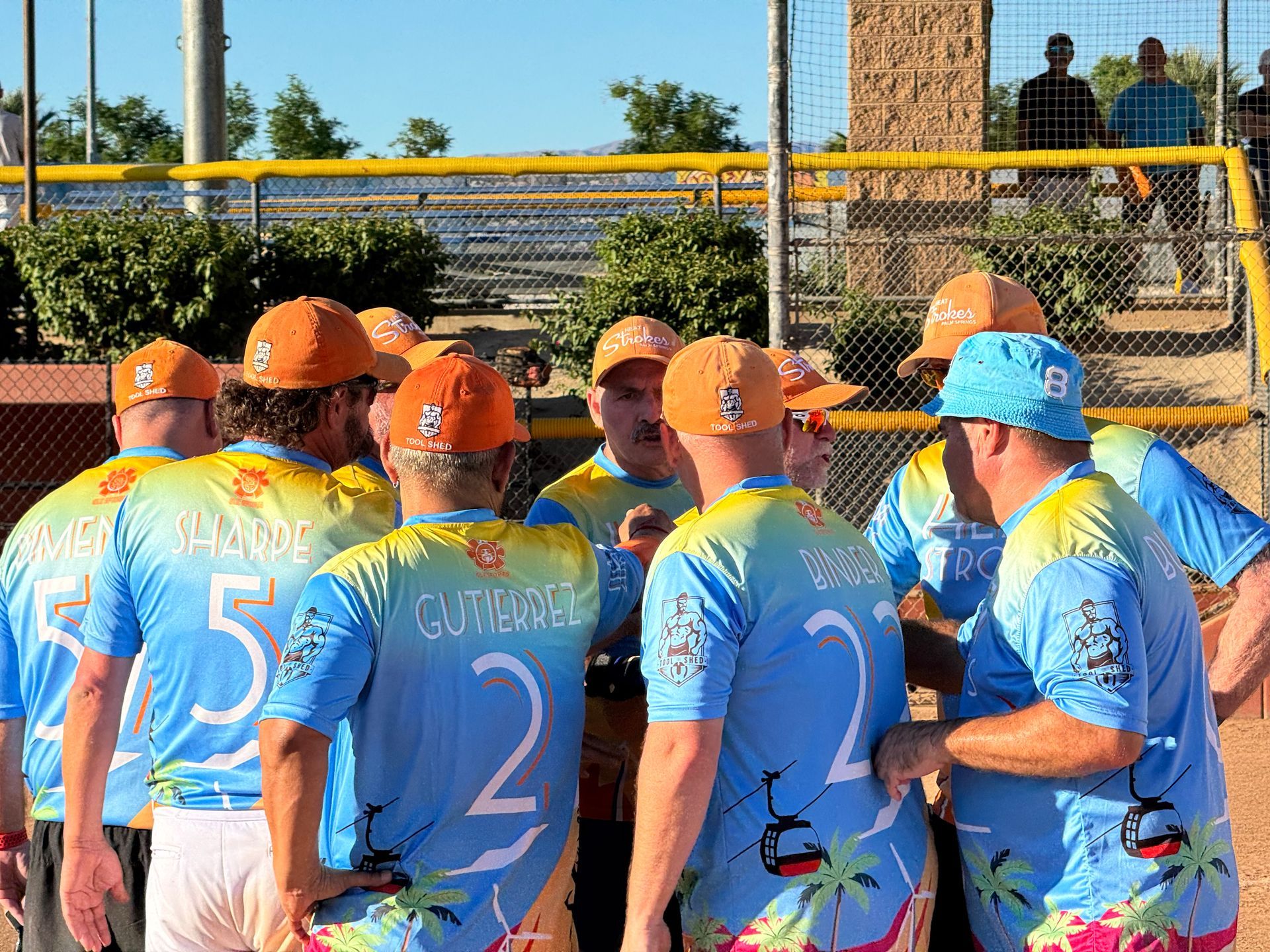 Softball team in blue and orange huddles near a yellow fence on the field