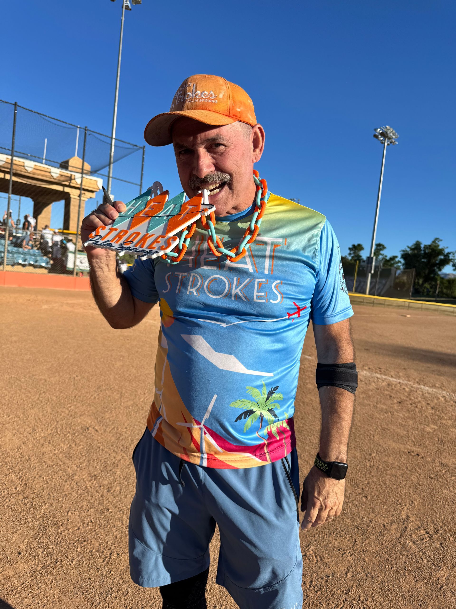 Man smiling in orange cap and blue shirt holding a fish on a sunny baseball field