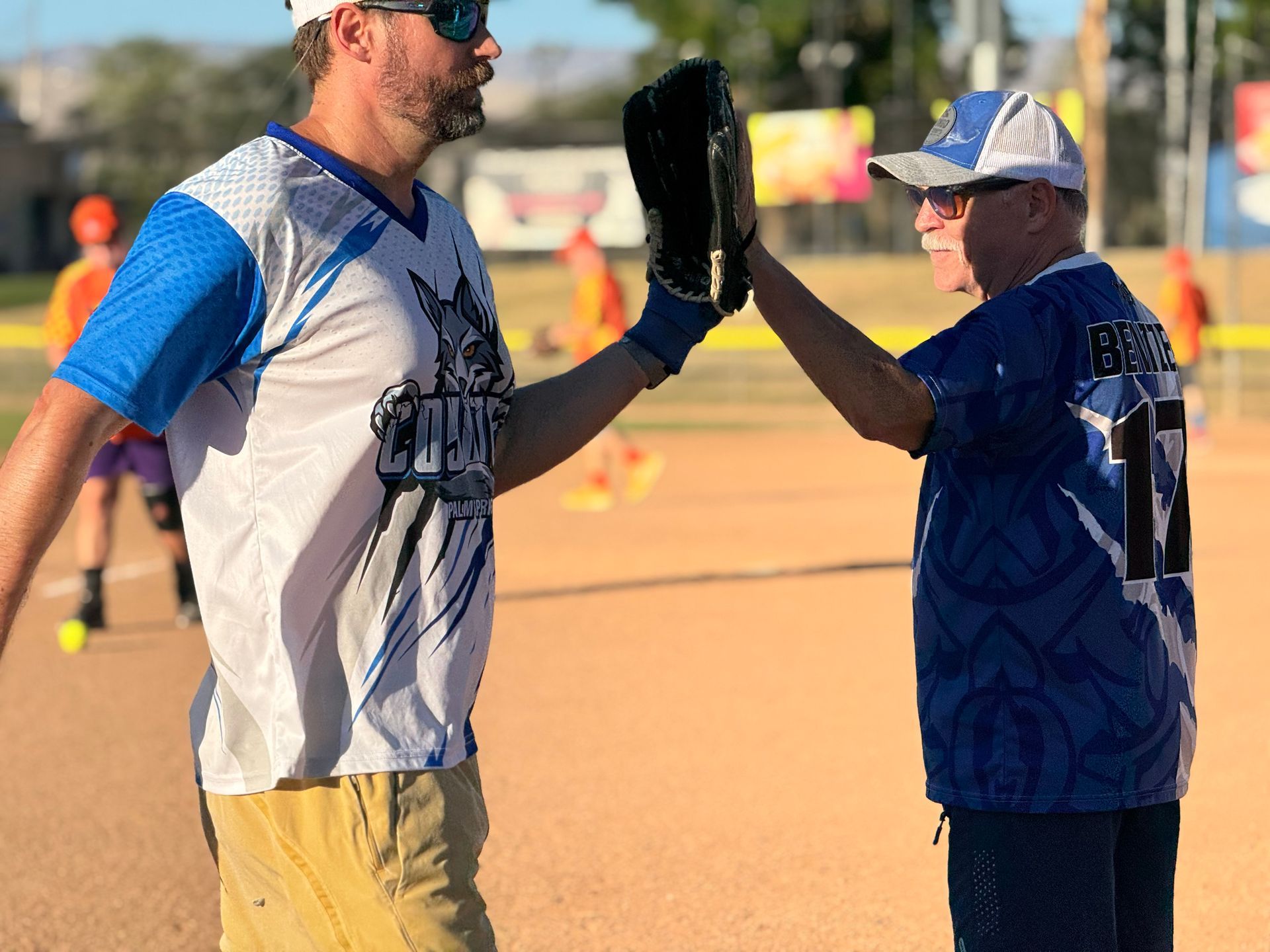 Two softball players high-five on a sunny field, one in gray and blue, the other in dark blue.