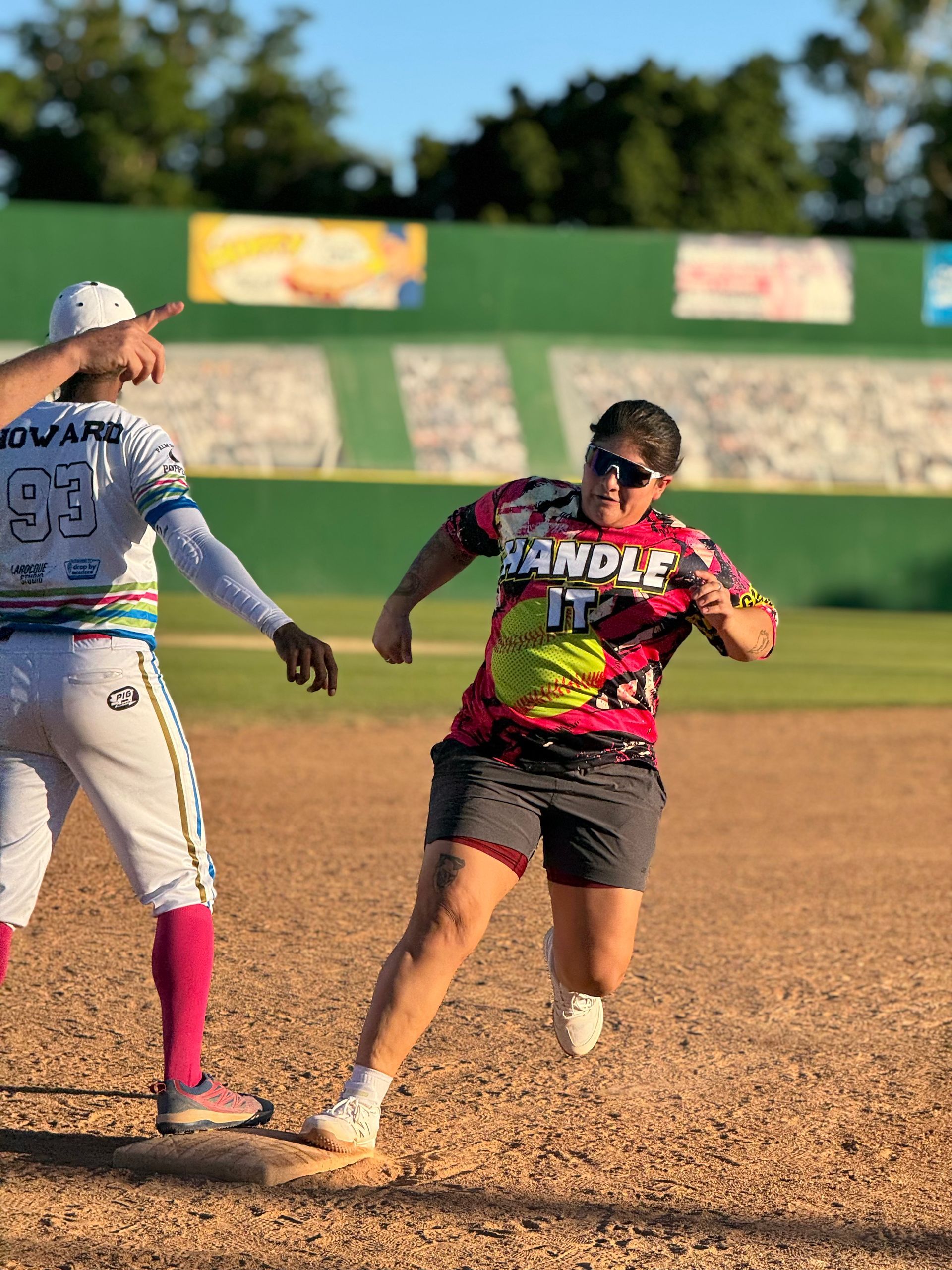 Two softball players sprinting on a dirt field, one in a pink-and-black jersey leading the play.