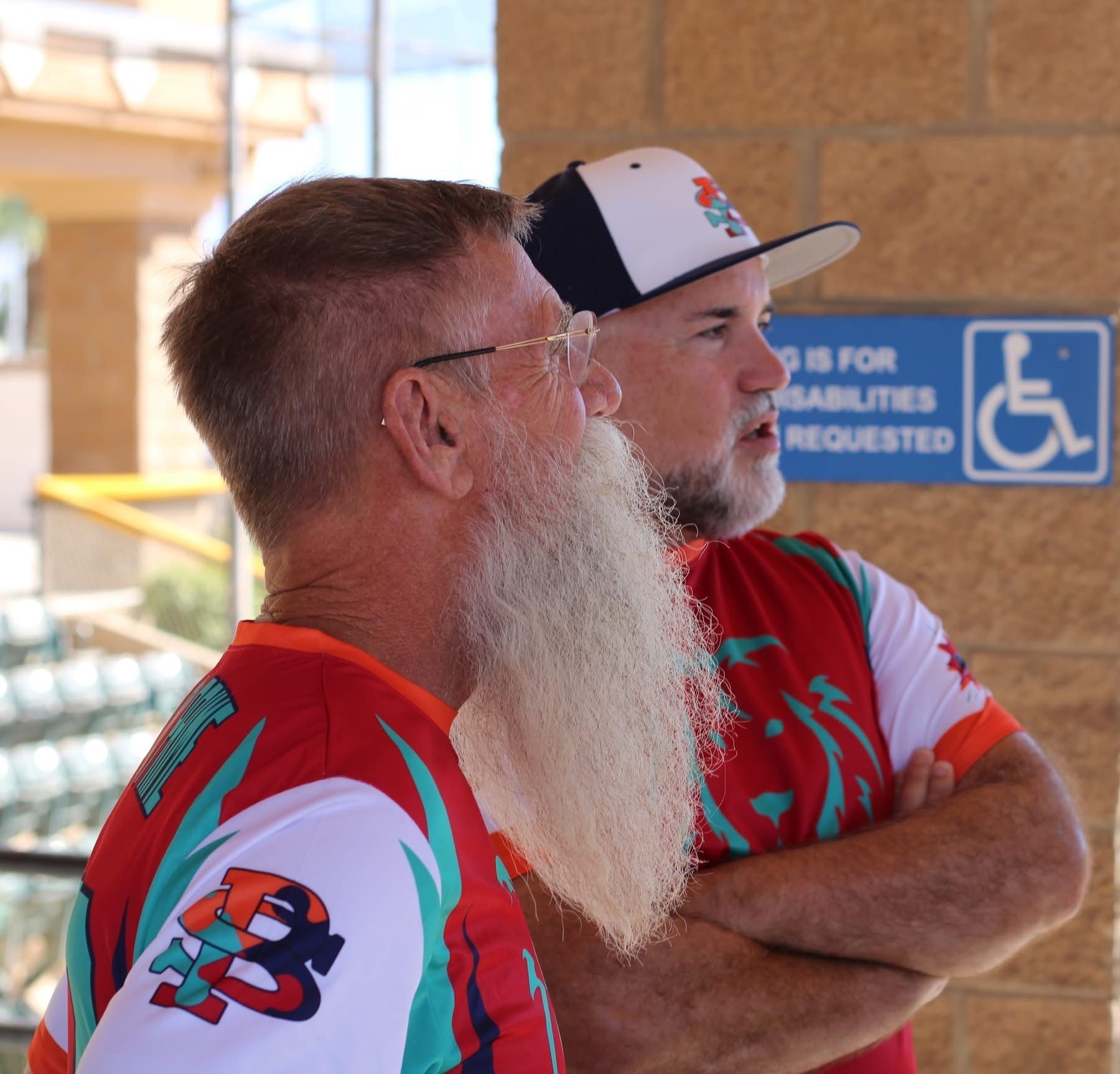 Two men in red shirts and caps standing outdoors, one with a long white beard, near a wheelchair-accessible sign