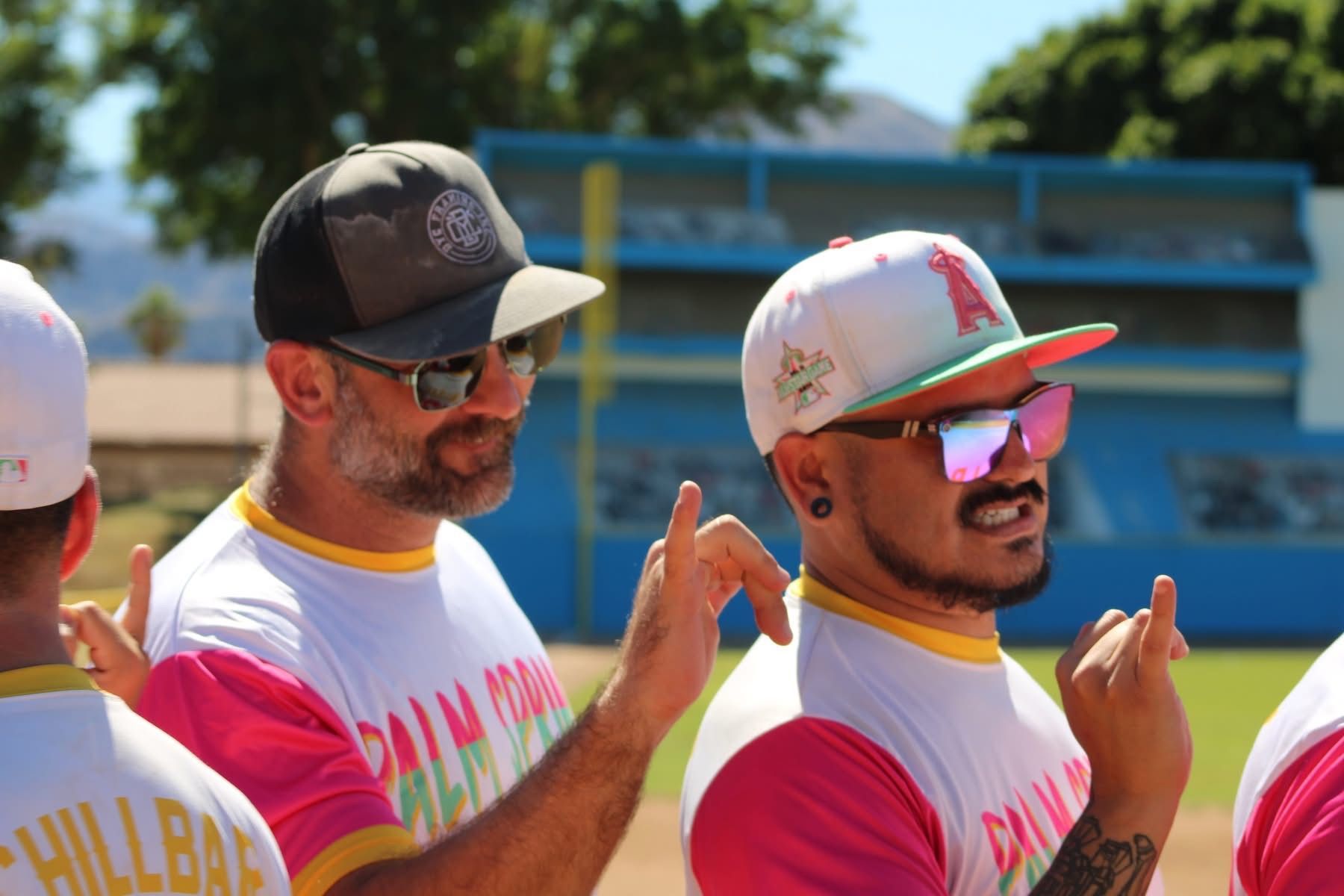 Two men in pink-and-white shirts and caps smiling, with one raising a finger at an outdoor event