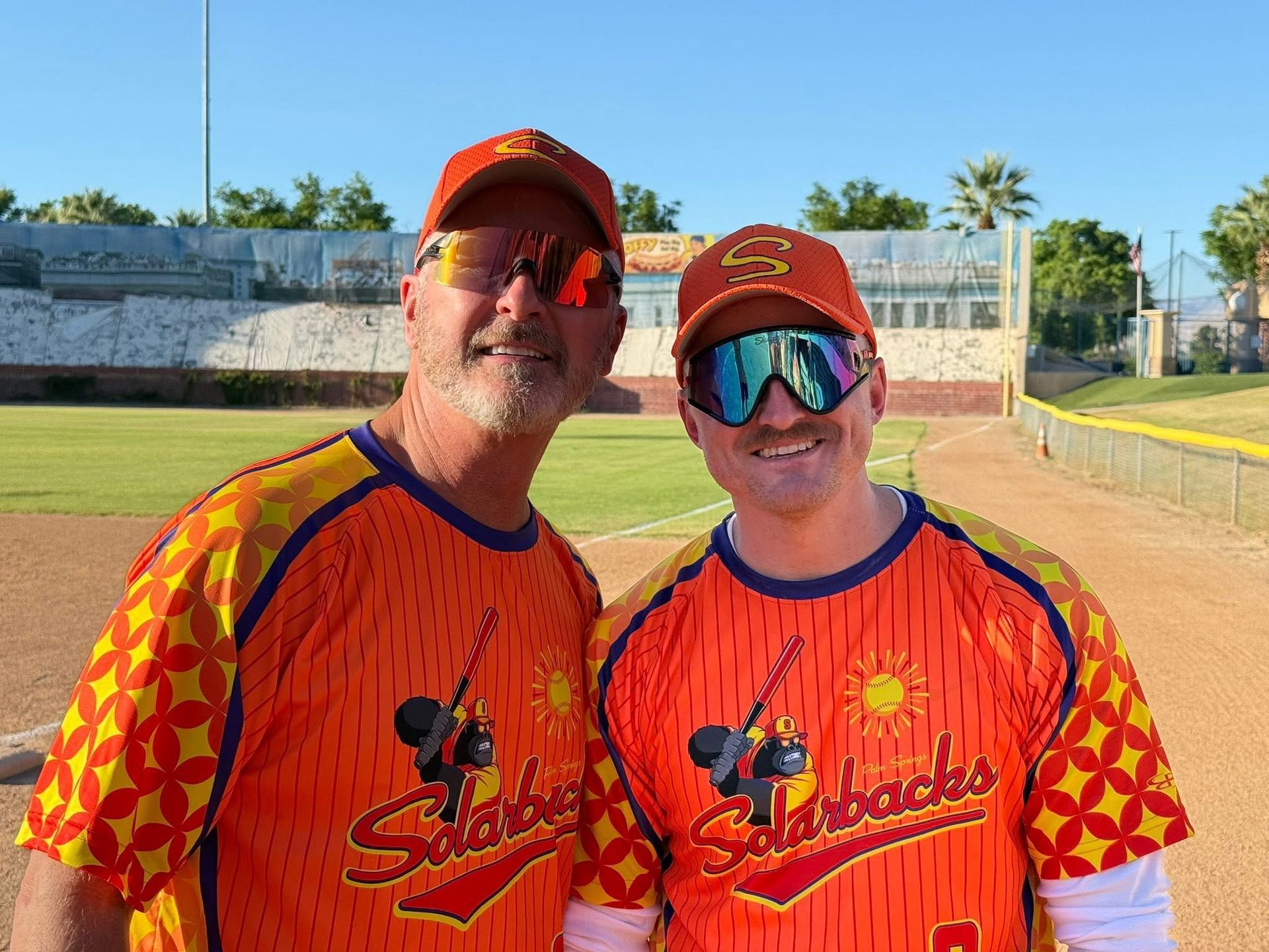 Two smiling teammates in orange jerseys and caps standing on a baseball field