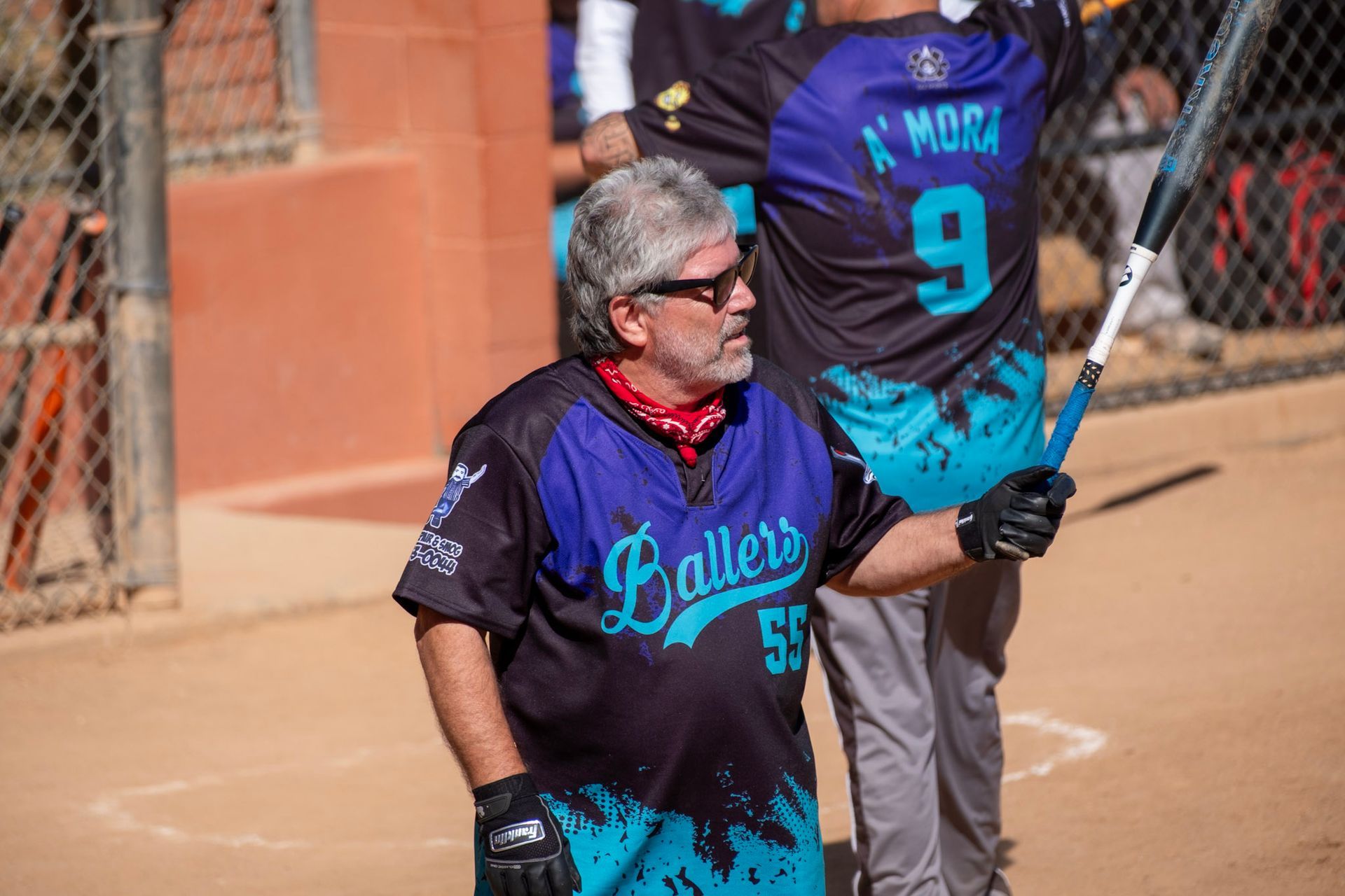 Softball player in black and blue uniform stands on dirt field, holding a bat near a chain-link fence.