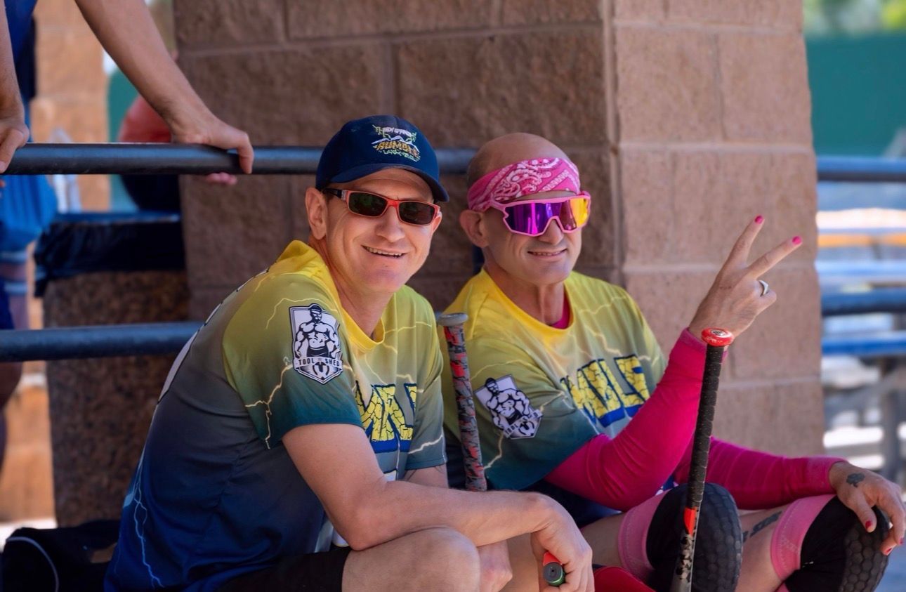 Two cyclists in yellow jerseys sit smiling on a bench, one pointing and wearing pink sunglasses and a headscarf.