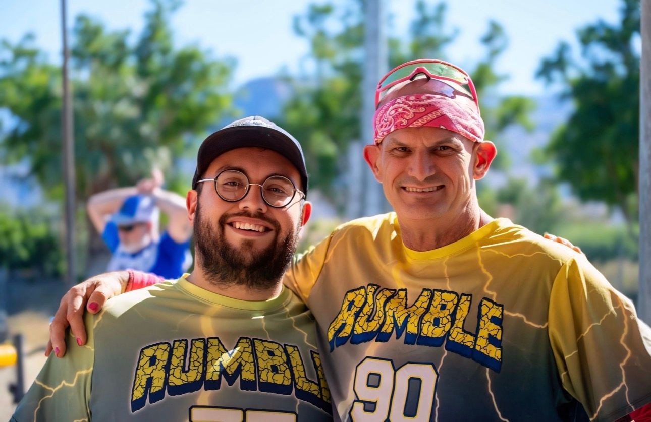 Two smiling people in yellow sports jerseys pose outdoors, one with an arm around the other.