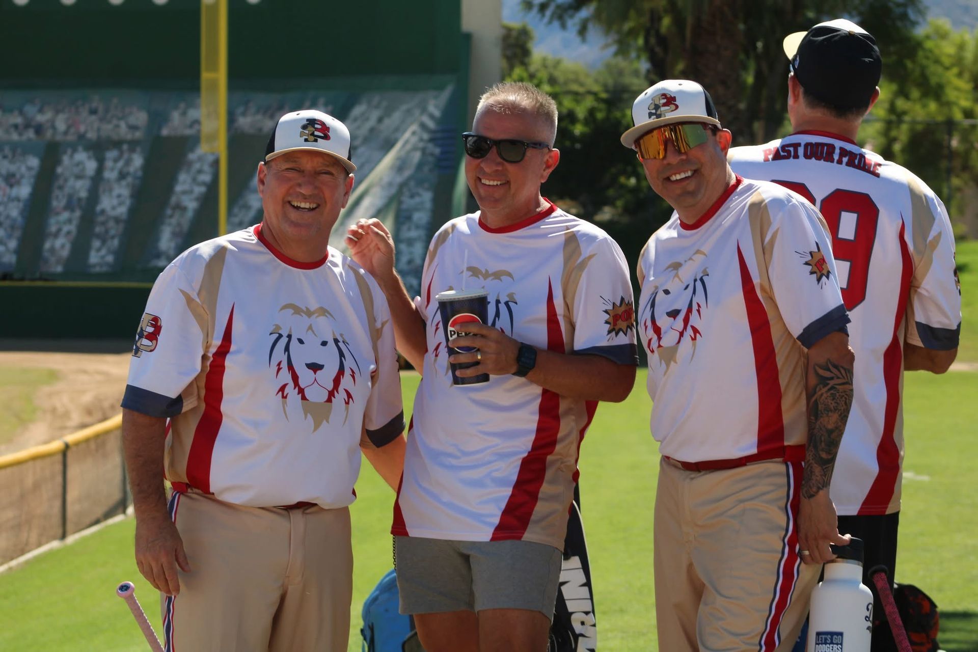 Four softball players in white and red jerseys smiling on a field, with one holding a drink杯