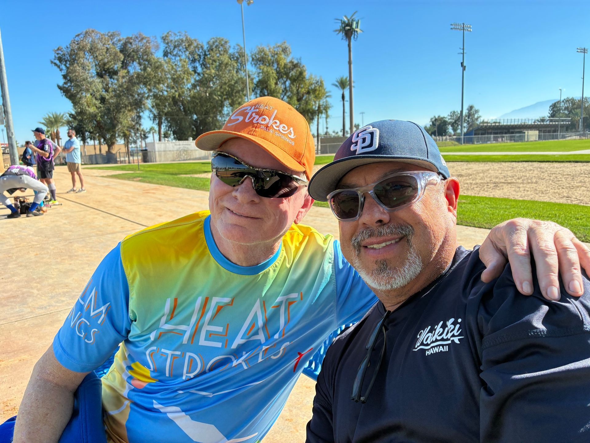 Two smiling friends in sunglasses and caps posing outdoors on a sunny field.