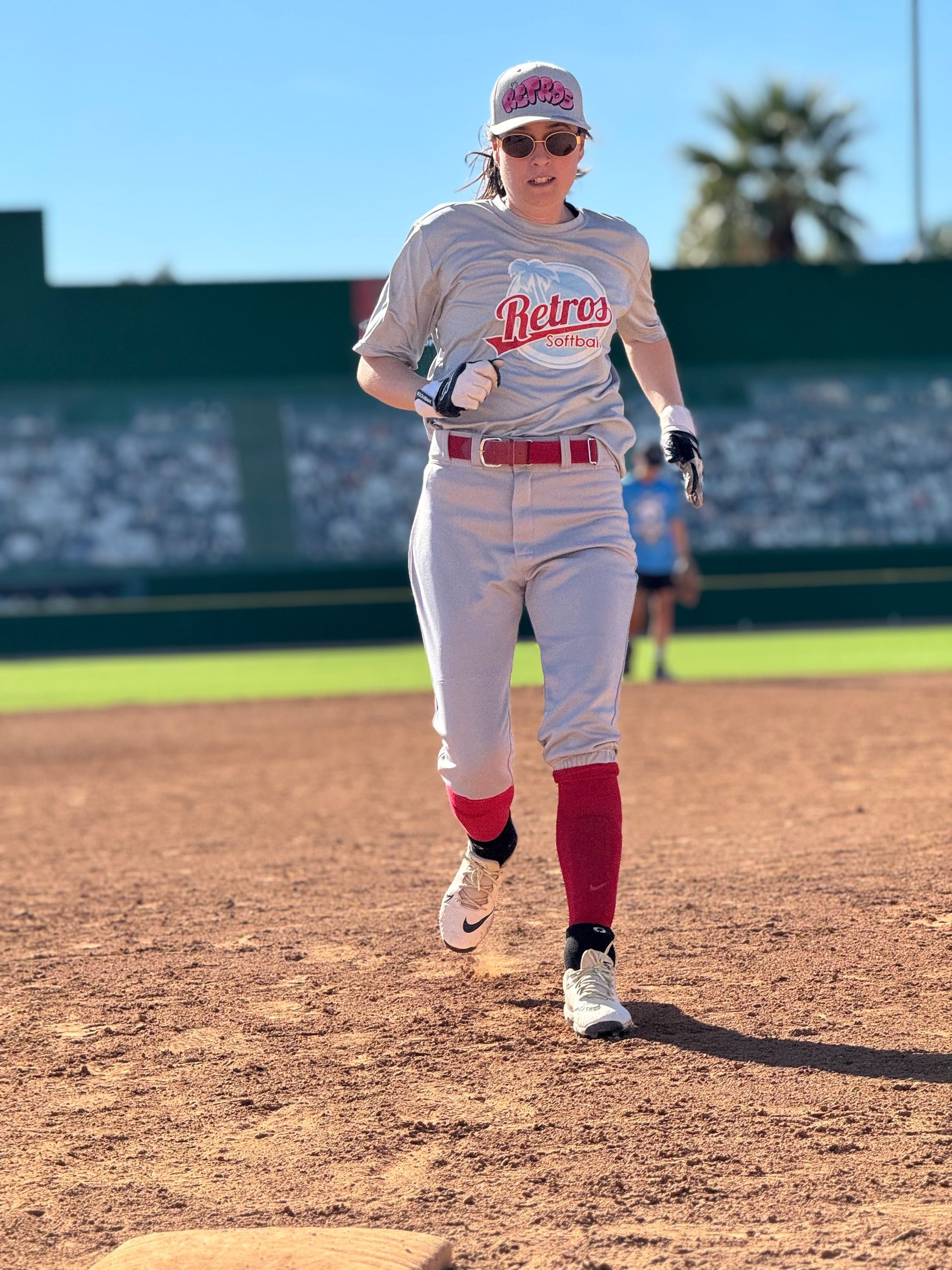 Softball player in gray and red uniform running on a dirt field under a blue sky.