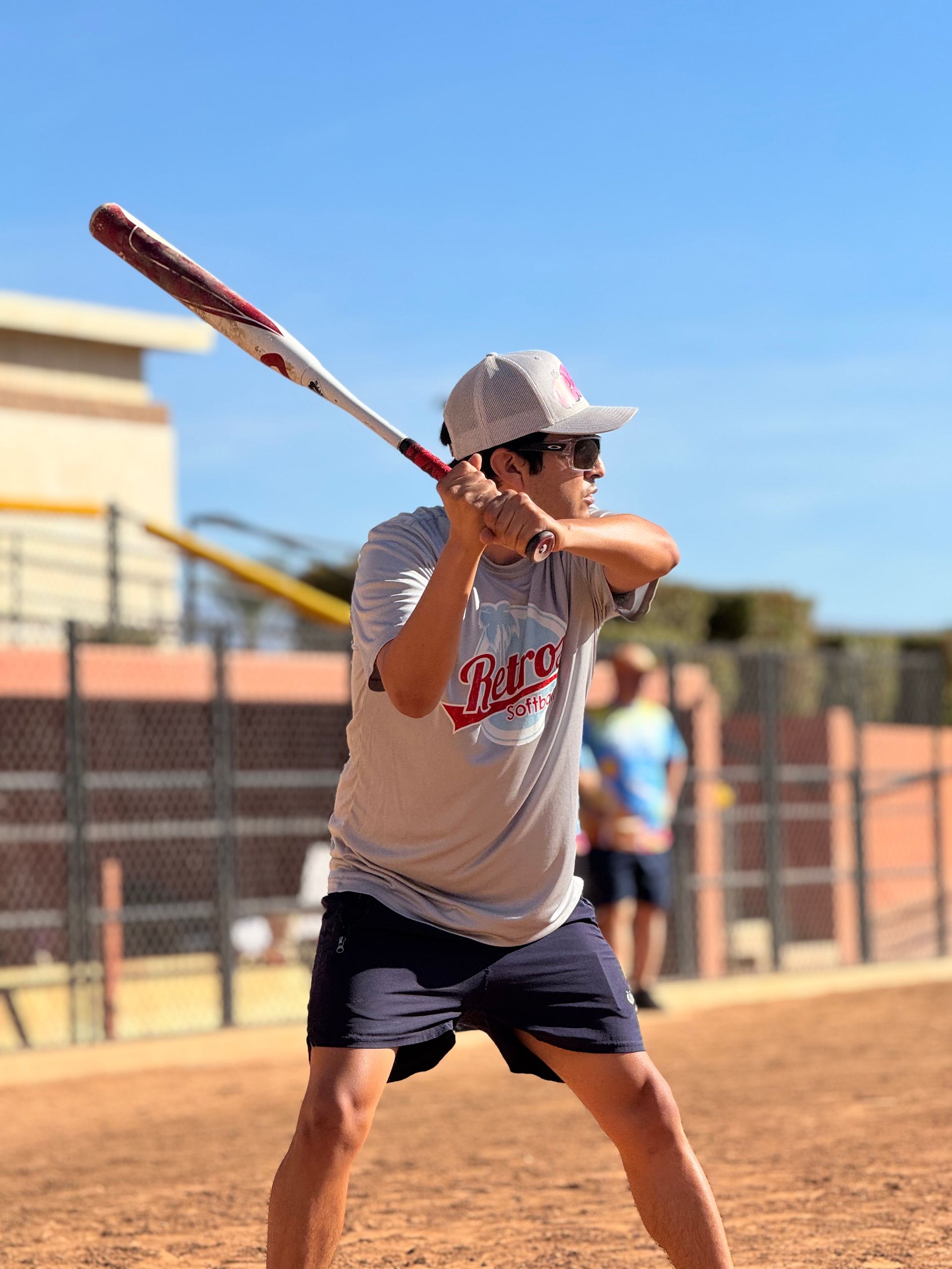 Baseball player in gray shirt and black shorts batting at a field under a clear sky