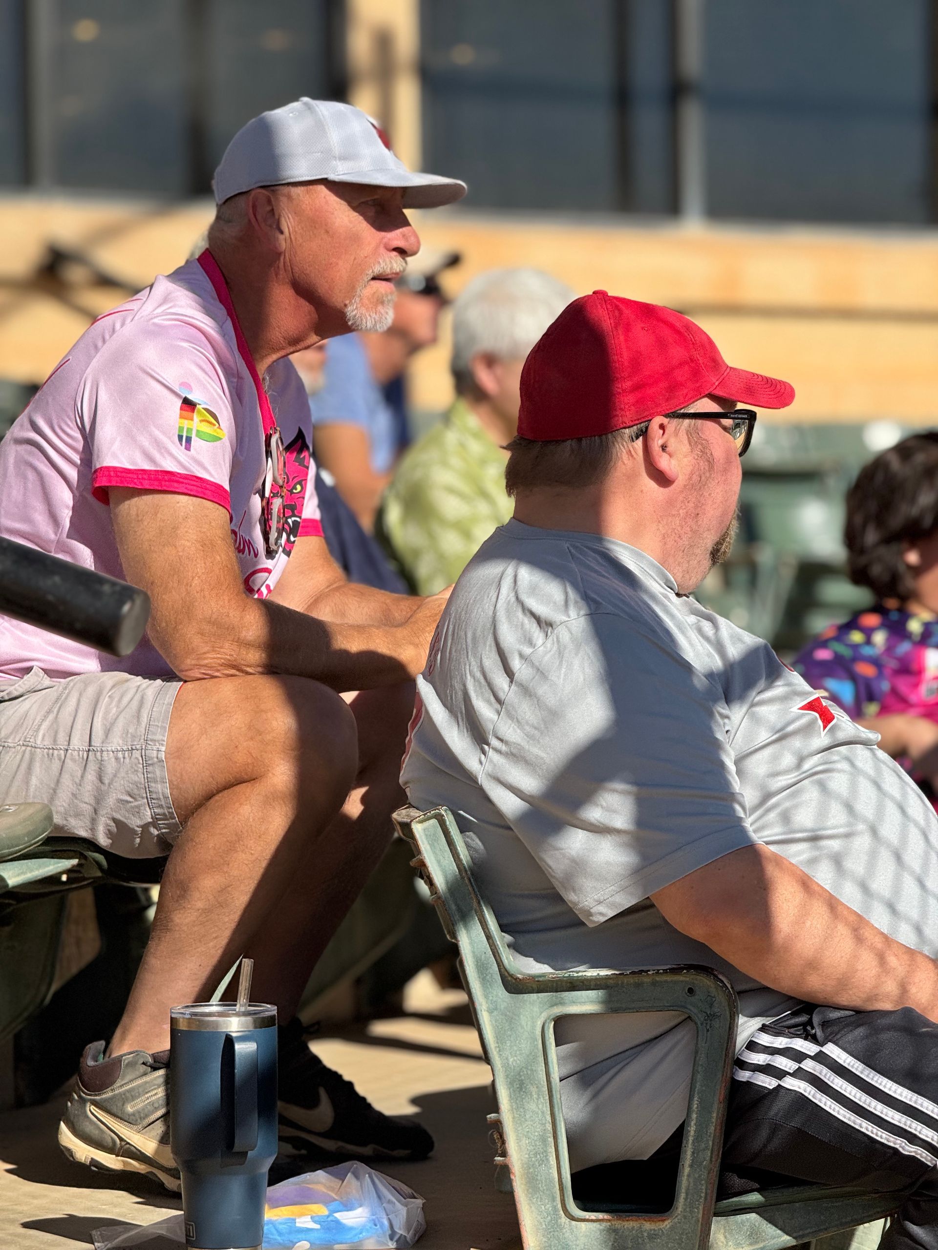 Spectators seated outdoors in sunlight, wearing caps and casual clothes, watching an event.