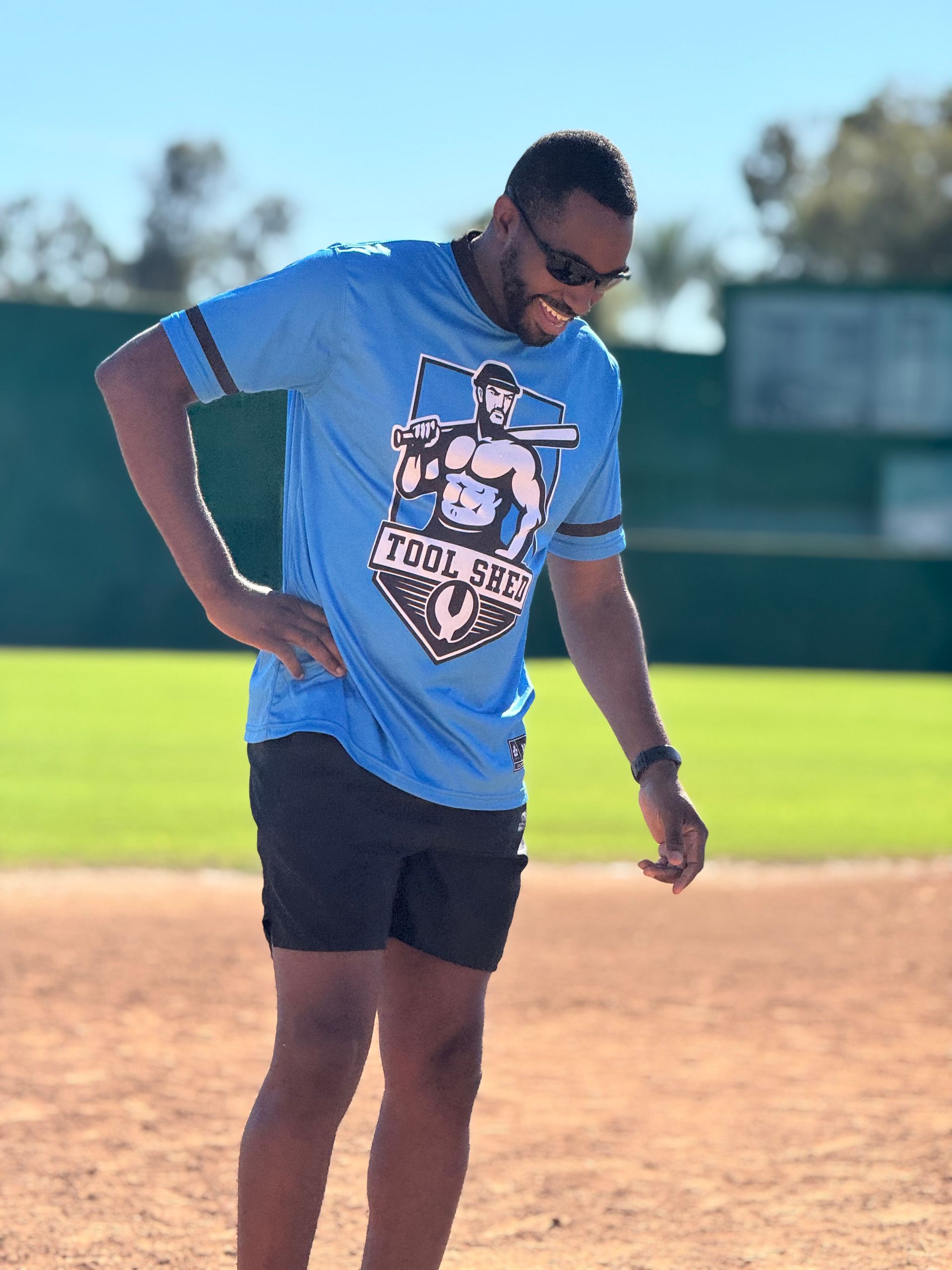 Man in a blue T-shirt and black shorts standing on a baseball field, looking down.