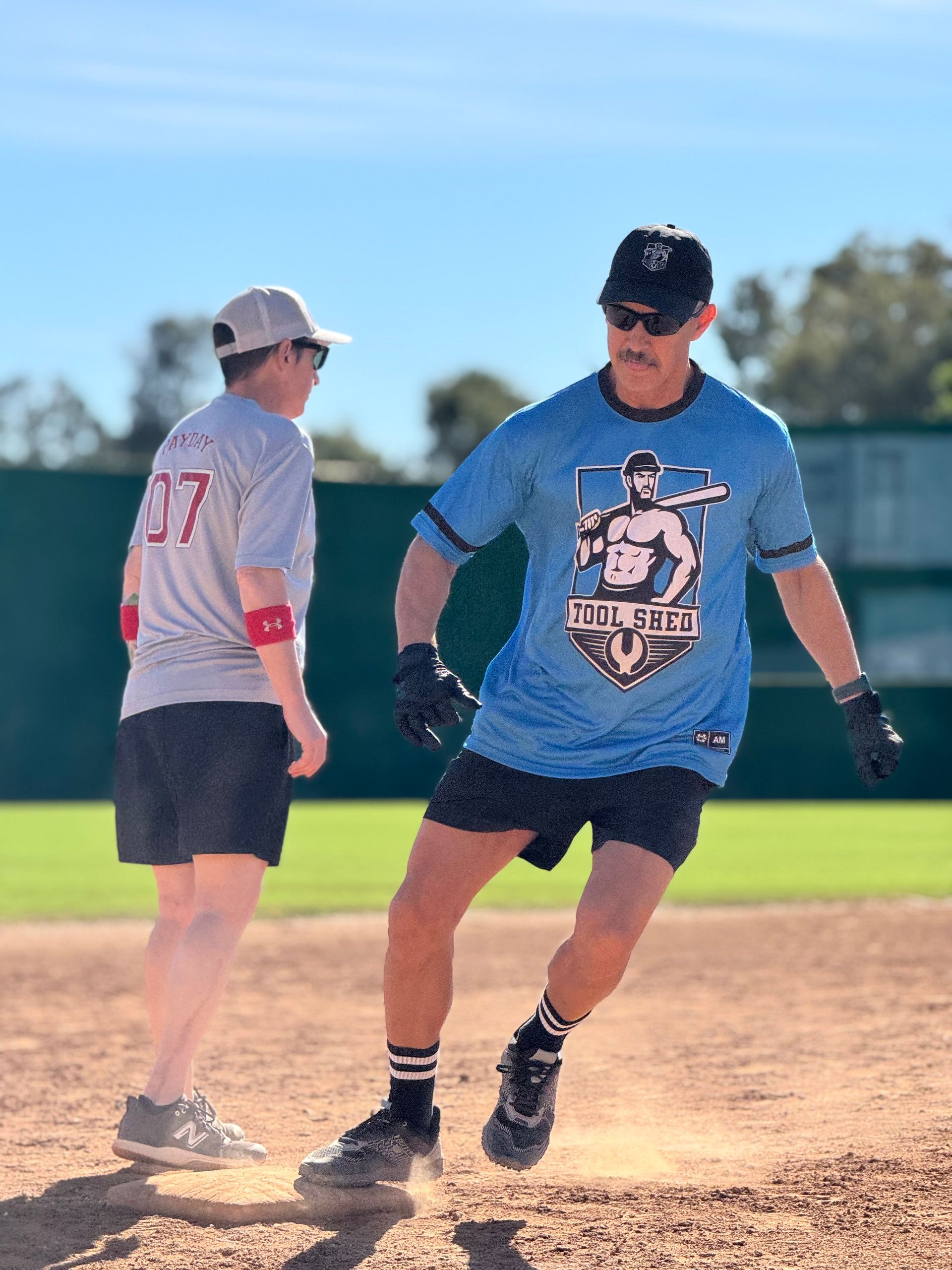 Two softball players on a dirt field, one in a blue shirt running past another in gray