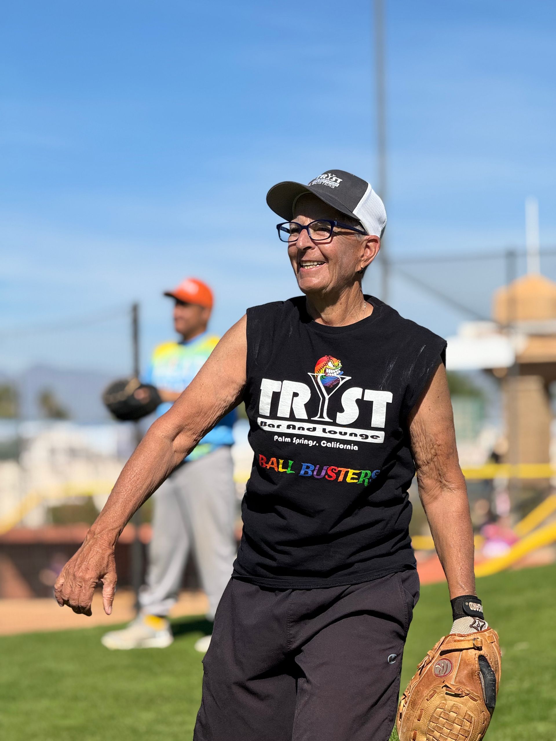 Smiling softball player in black shirt and cap, holding glove on a sunny field