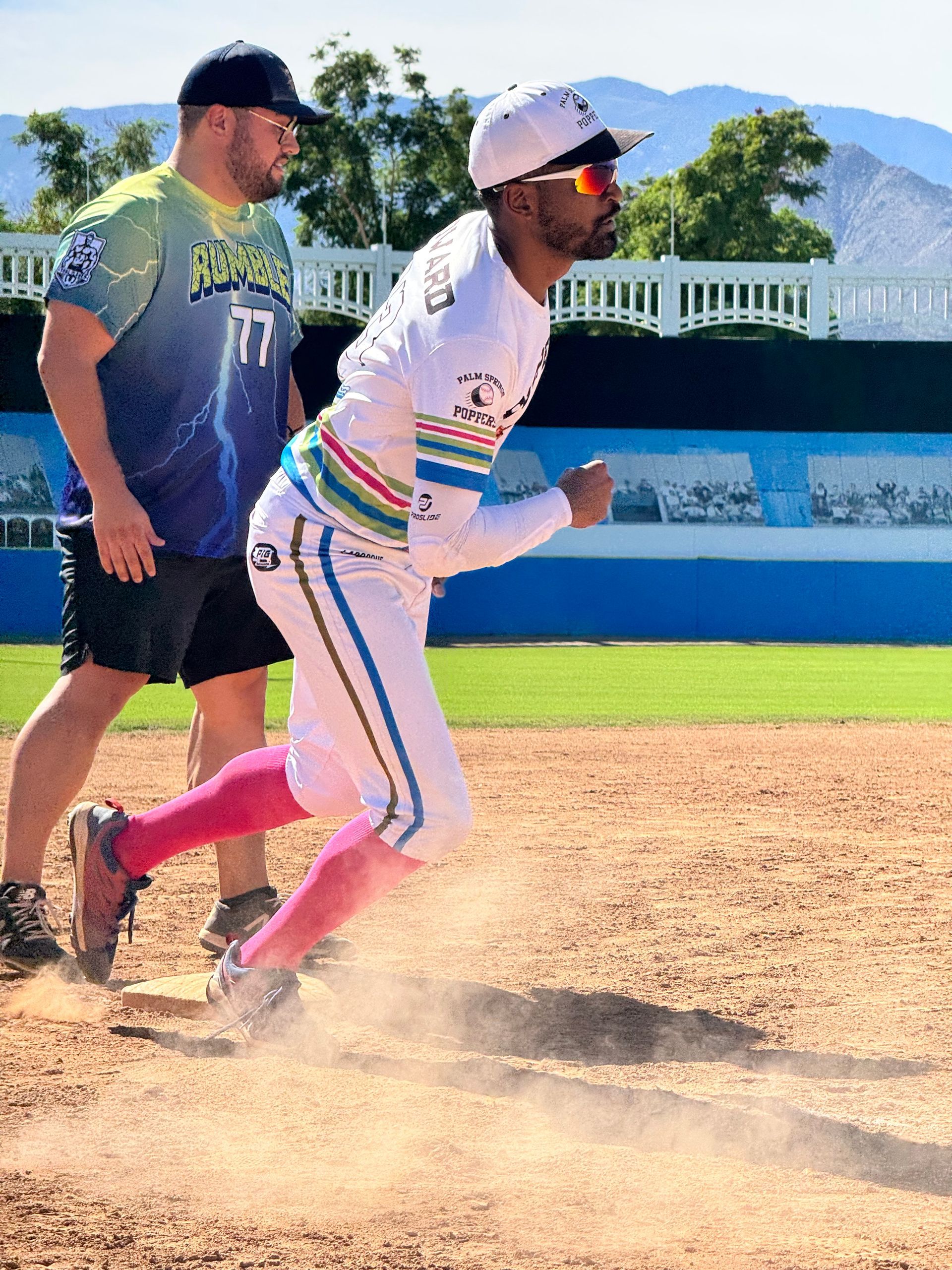 Two baseball players run the bases on a sunny field, with dust kicking up behind them.