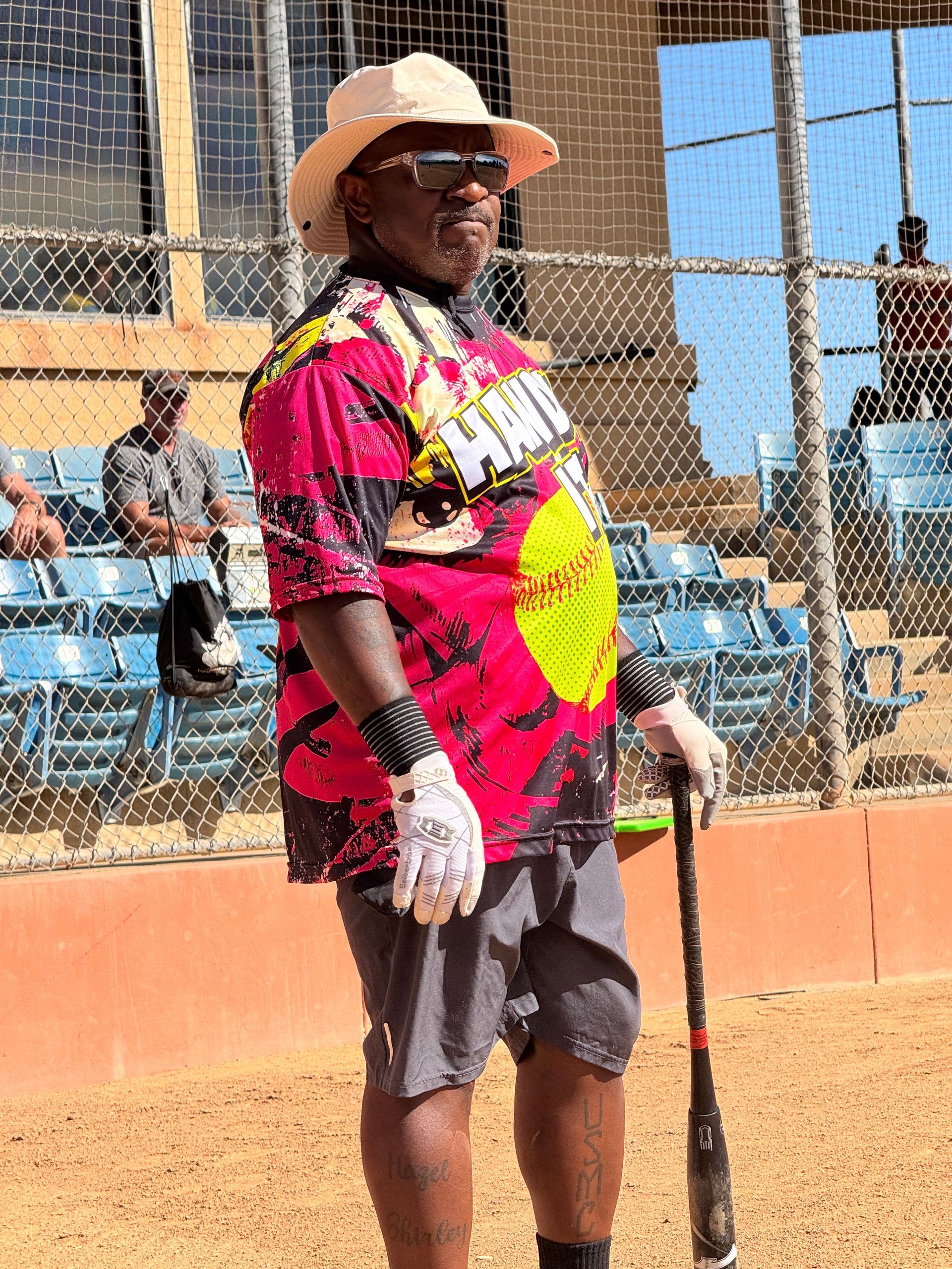 Person in a pink shirt, gray shorts, and straw hat stands with a bat near a softball field fence.