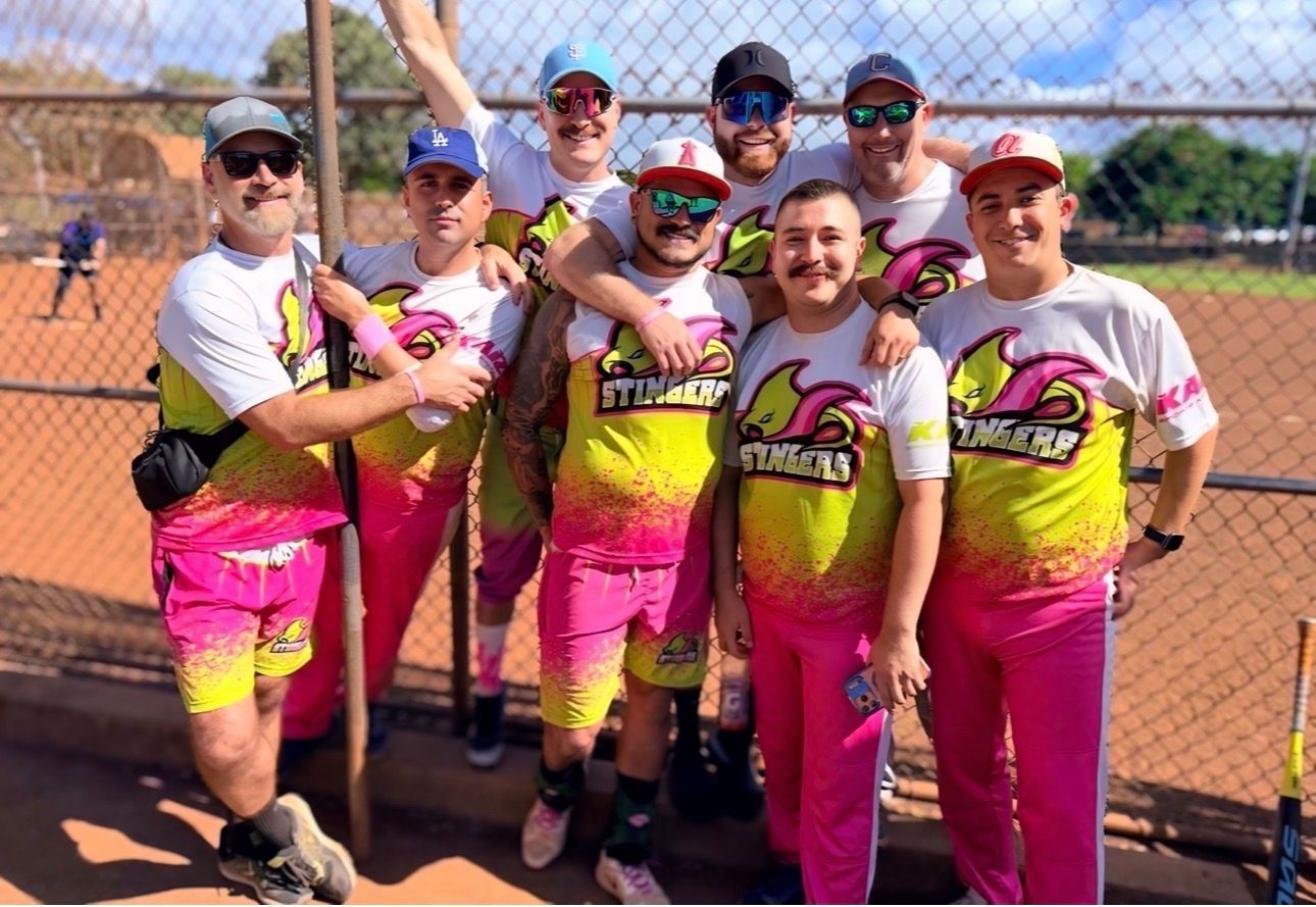 Softball team in matching pink and yellow uniforms posing together on a field behind a chain-link fence
