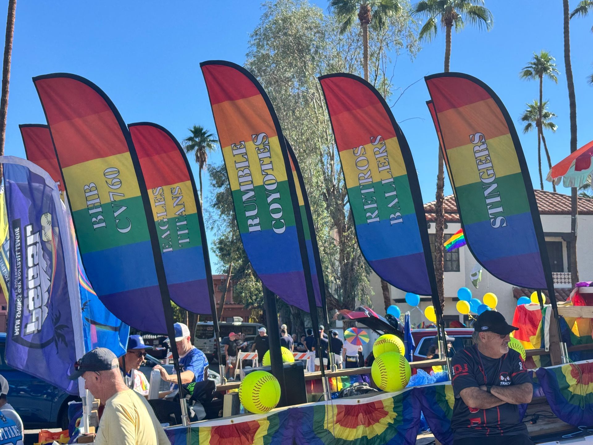 Rainbow flags at a sunny outdoor event with people, balloons, and palm trees in the background