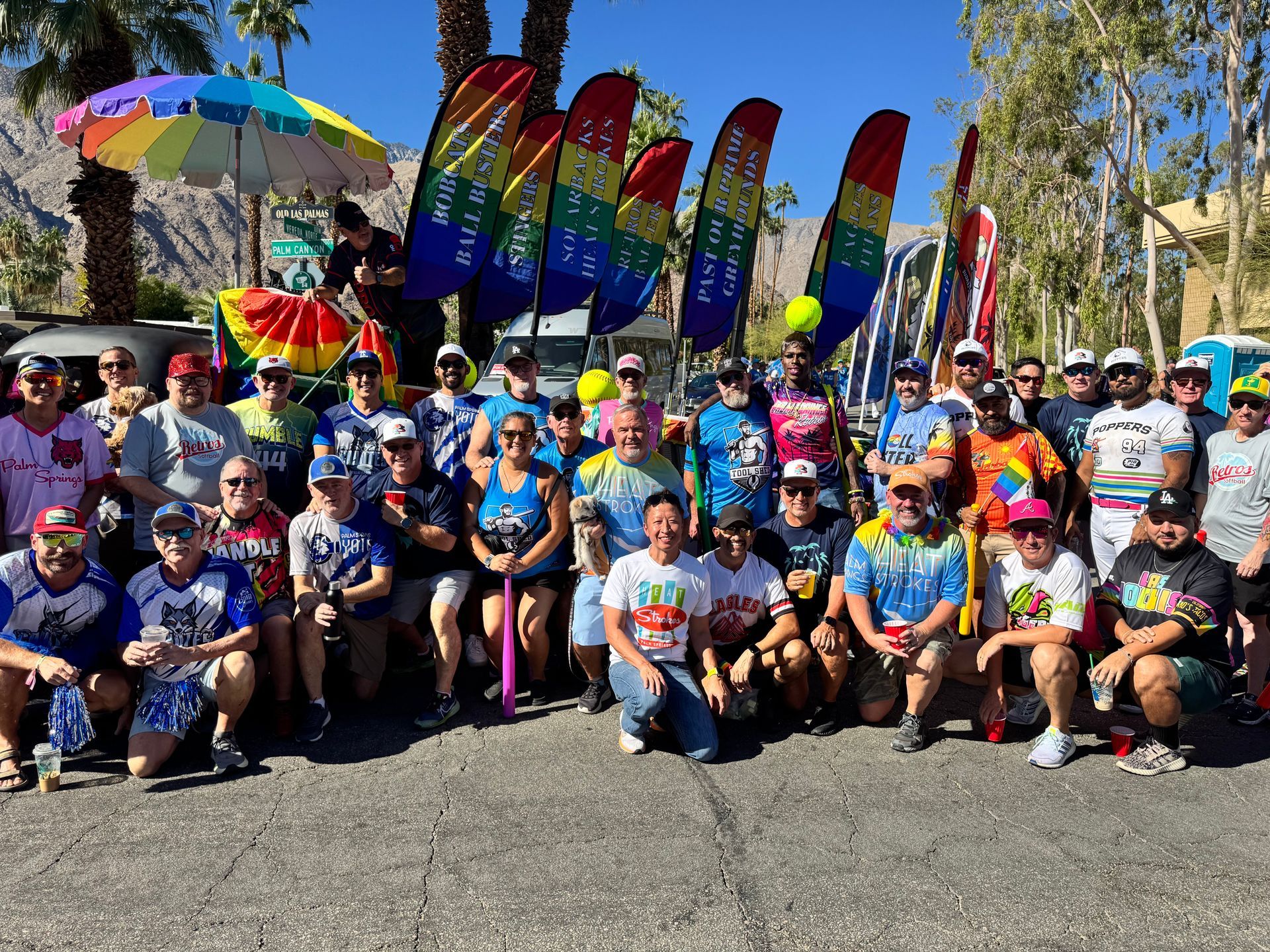 Large group posing outdoors with rainbow flags and colorful shirts at a pride event