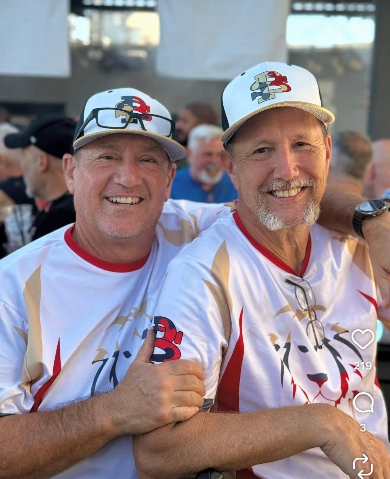 Two smiling men in matching white shirts and caps pose together outdoors.