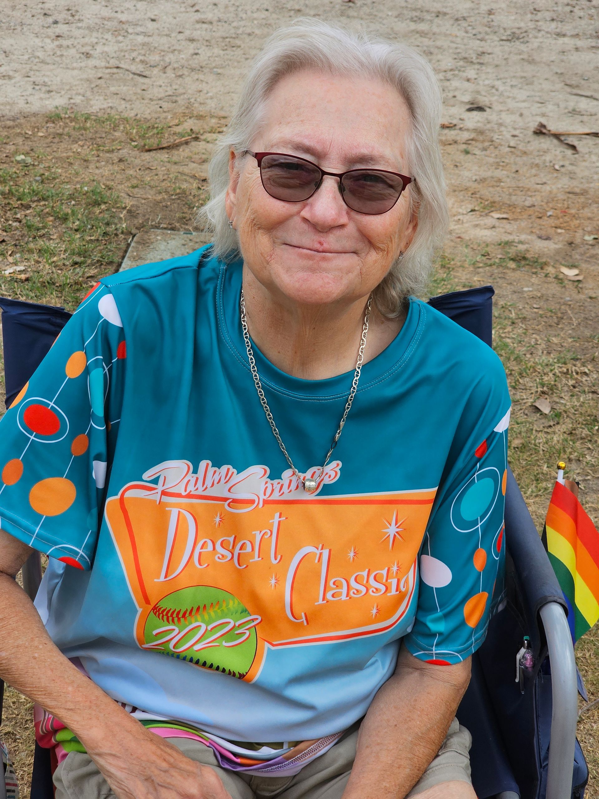 Smiling woman in sunglasses wearing a teal “Desert Oasis” shirt, seated outdoors in a chair.