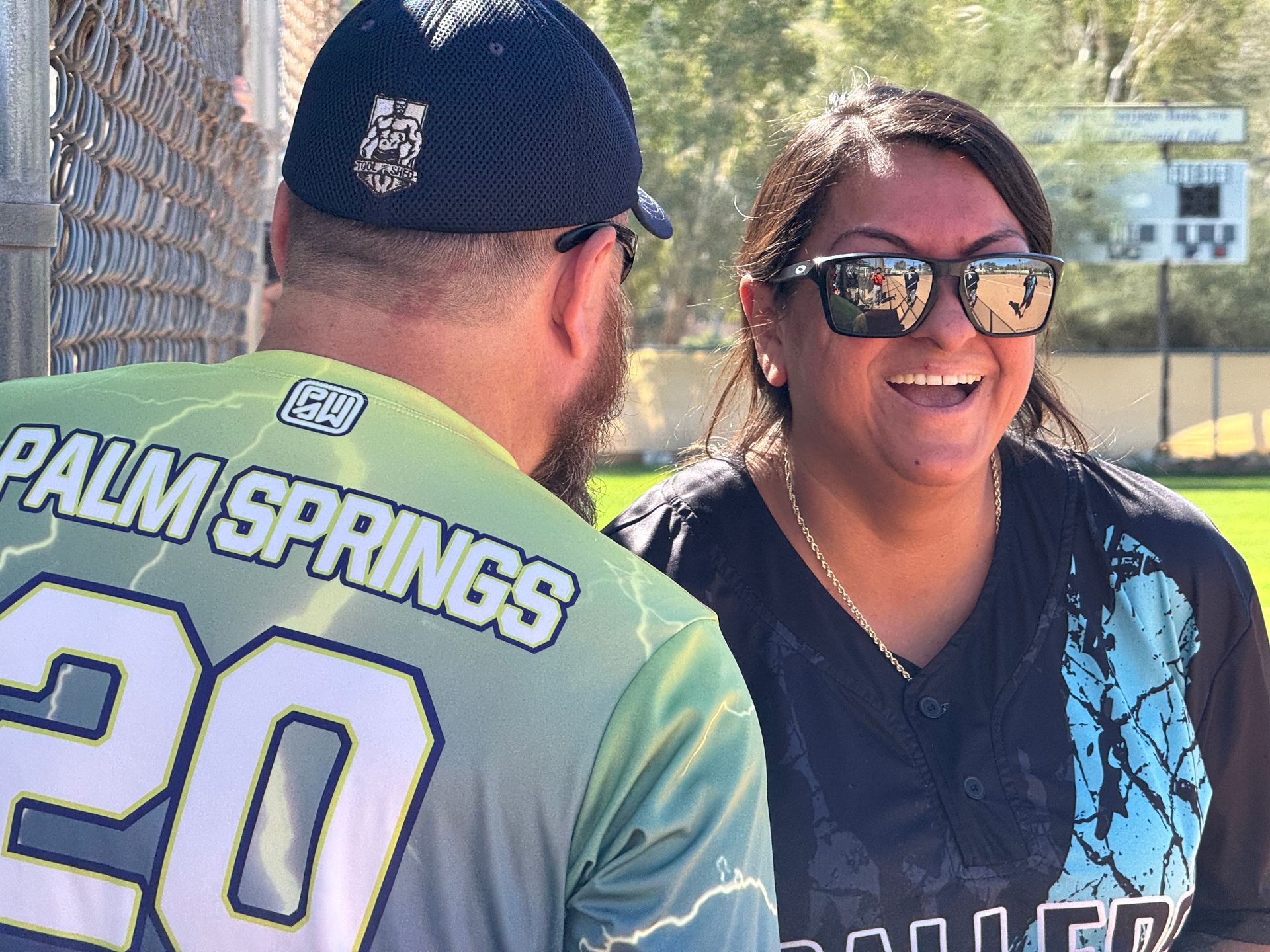 Two people smiling outdoors; one wears a Palm Springs jersey, the other sunglasses and a black jacket.
