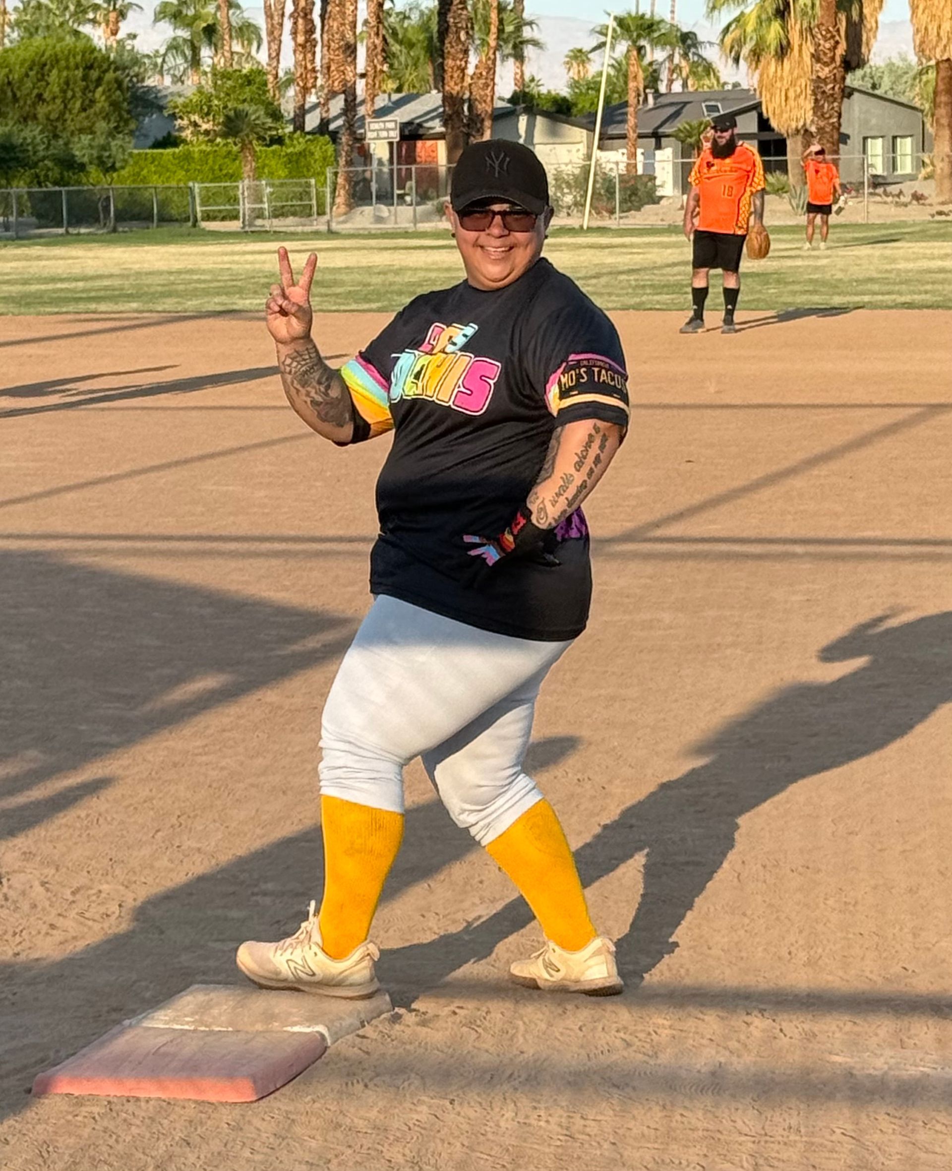Person in black shirt and gray leggings posing on a softball field, flashing a peace sign.