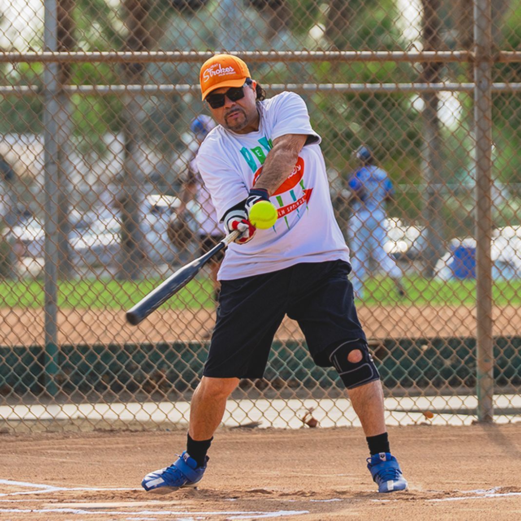 Softball player in orange cap swings a bat at a pitch on a field, with a chain-link fence behind him.