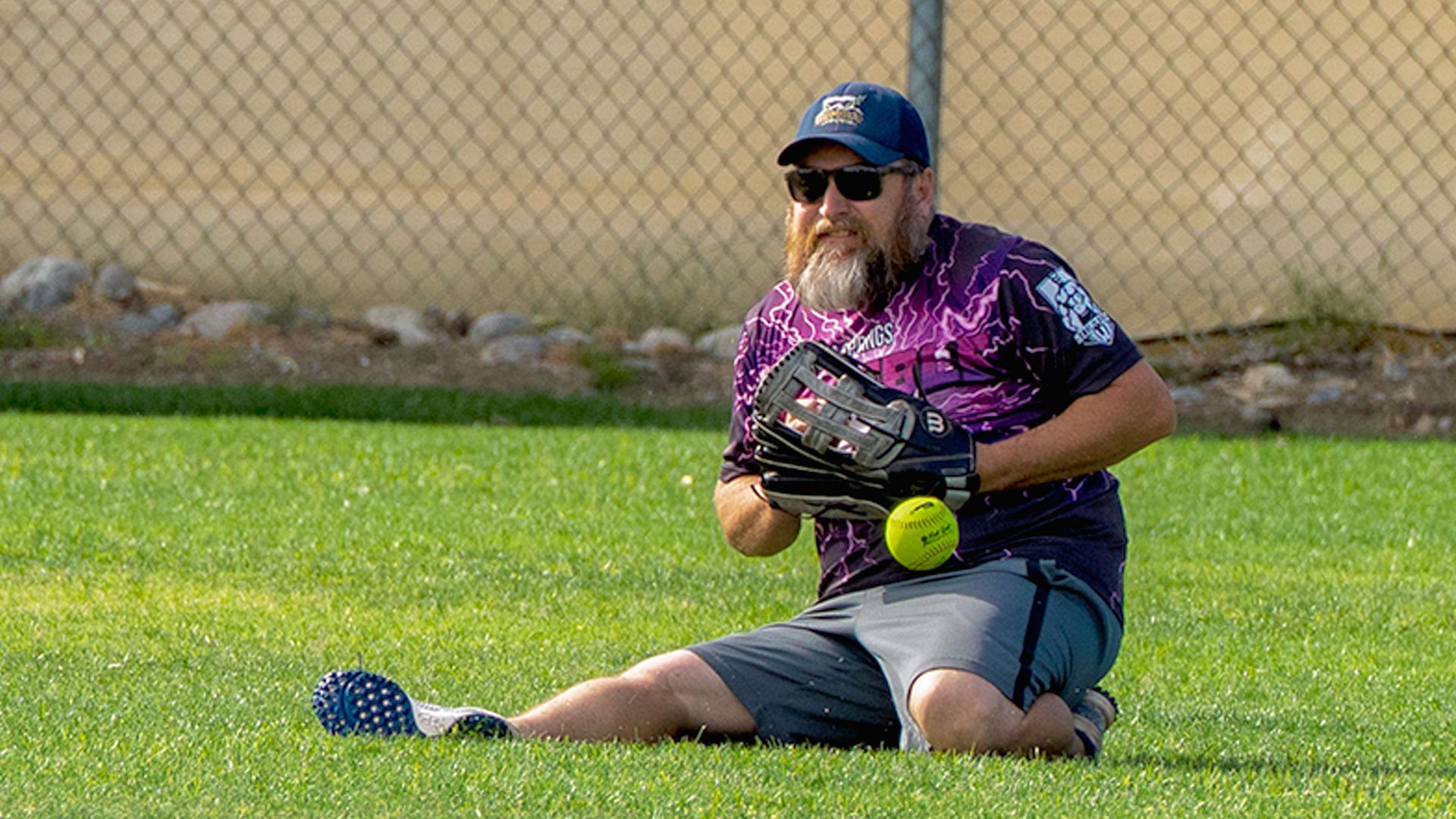 Man kneeling on grass, holding a softball glove and yellow ball near a chain-link fence.