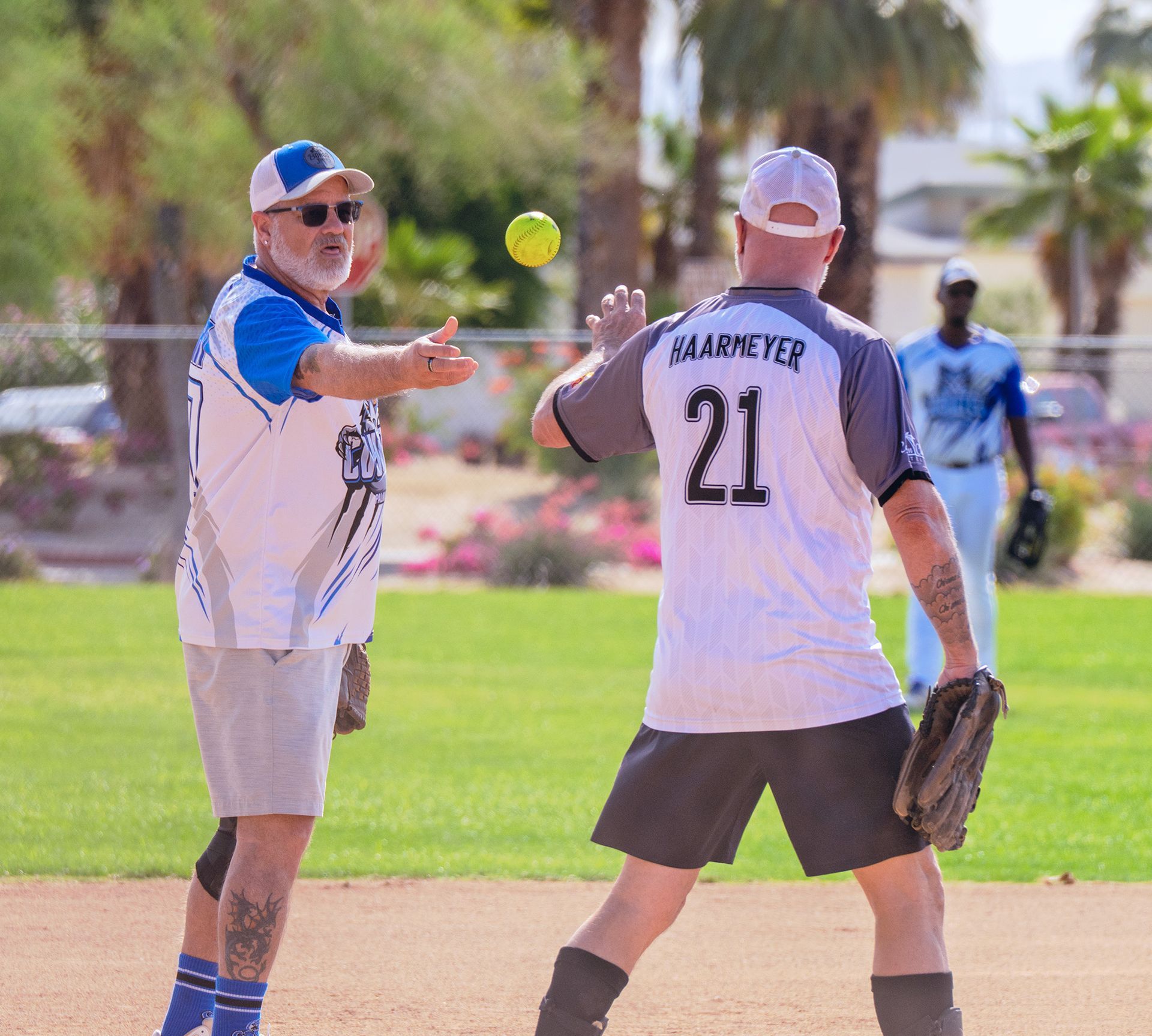 Two softball players on a field, one preparing to catch a yellow ball while a teammate watches.