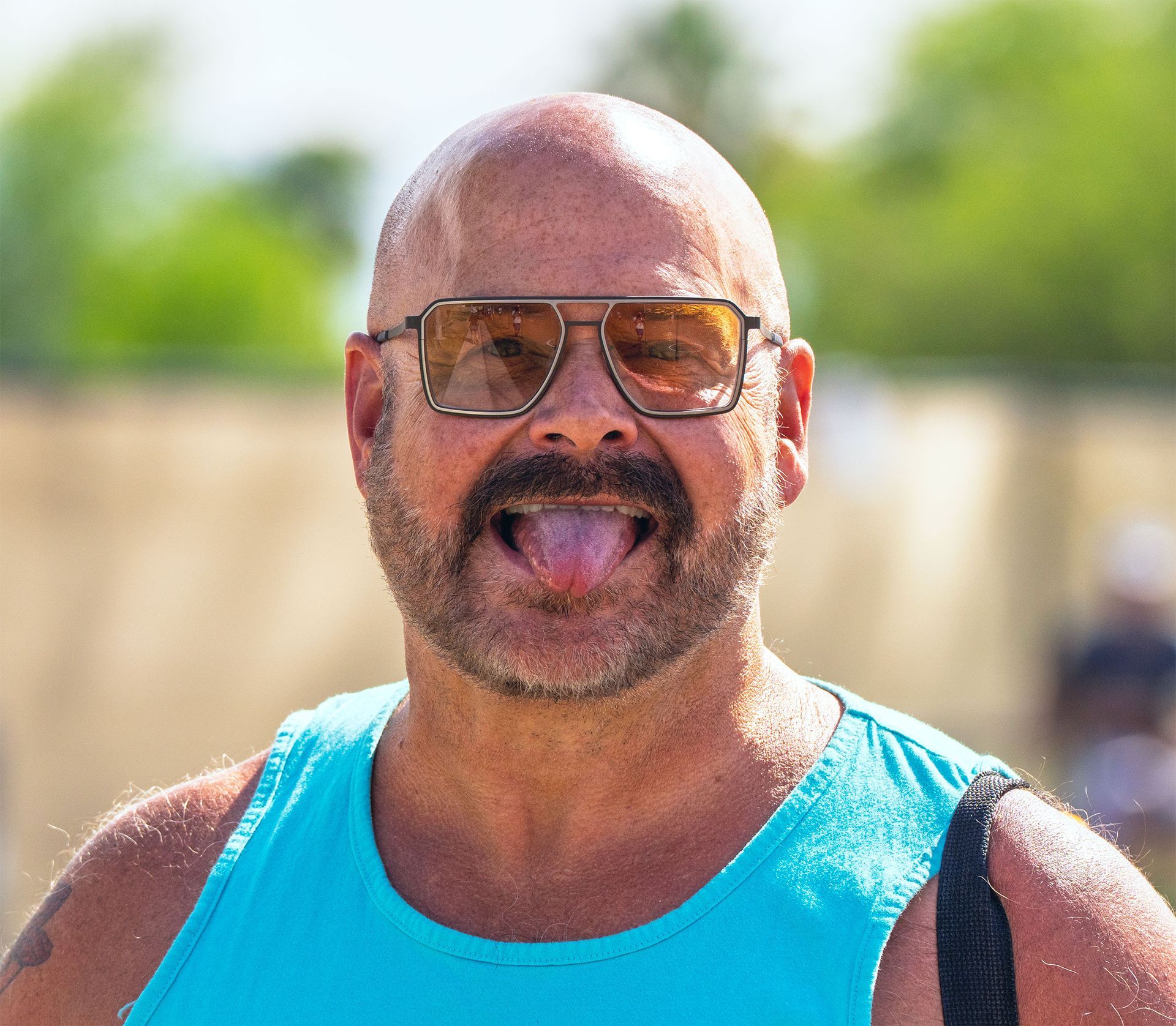 Man in sunglasses and turquoise tank top sticking out his tongue outdoors in bright sunlight