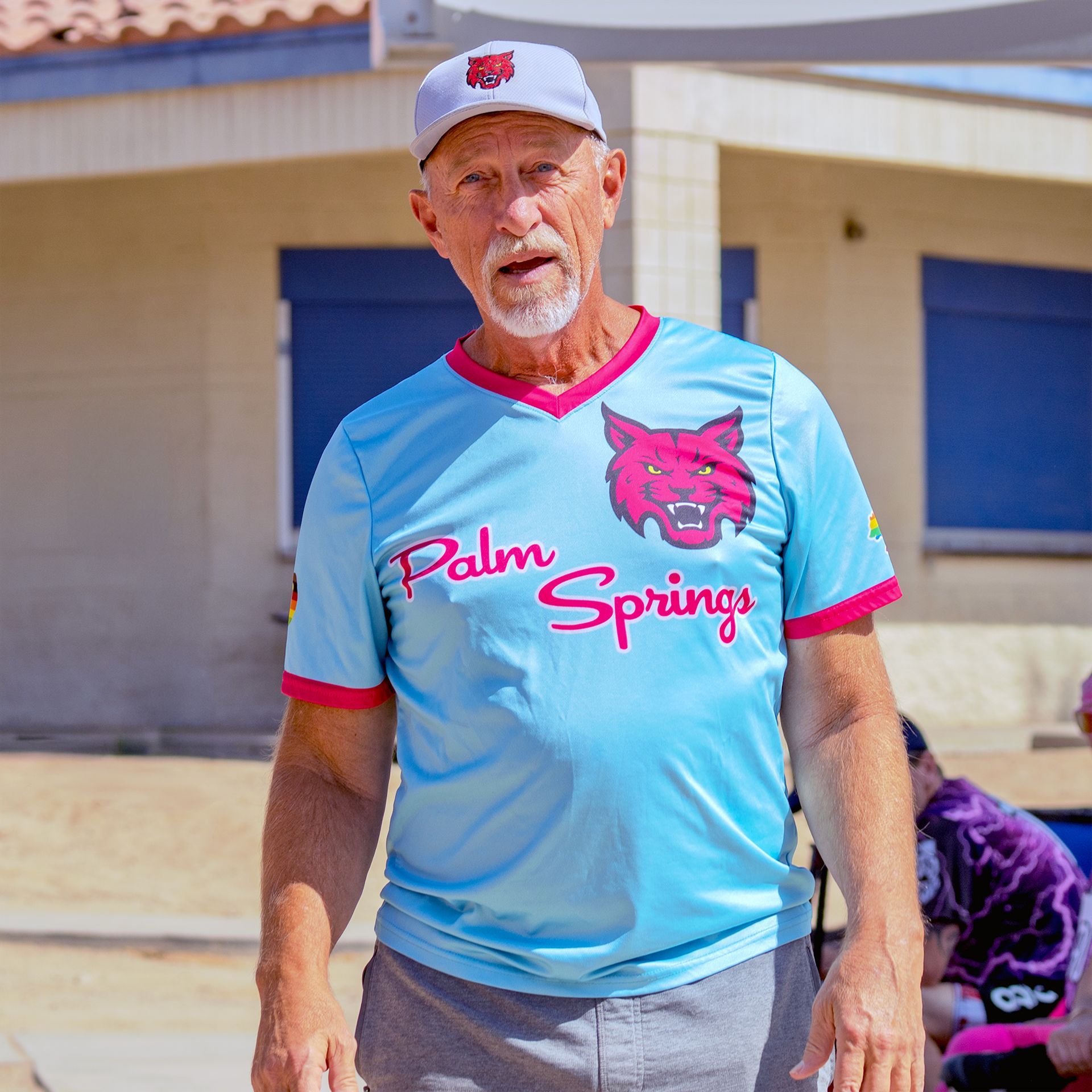 Man in a light blue Palm Springs jersey and cap standing outdoors by a building in bright sun