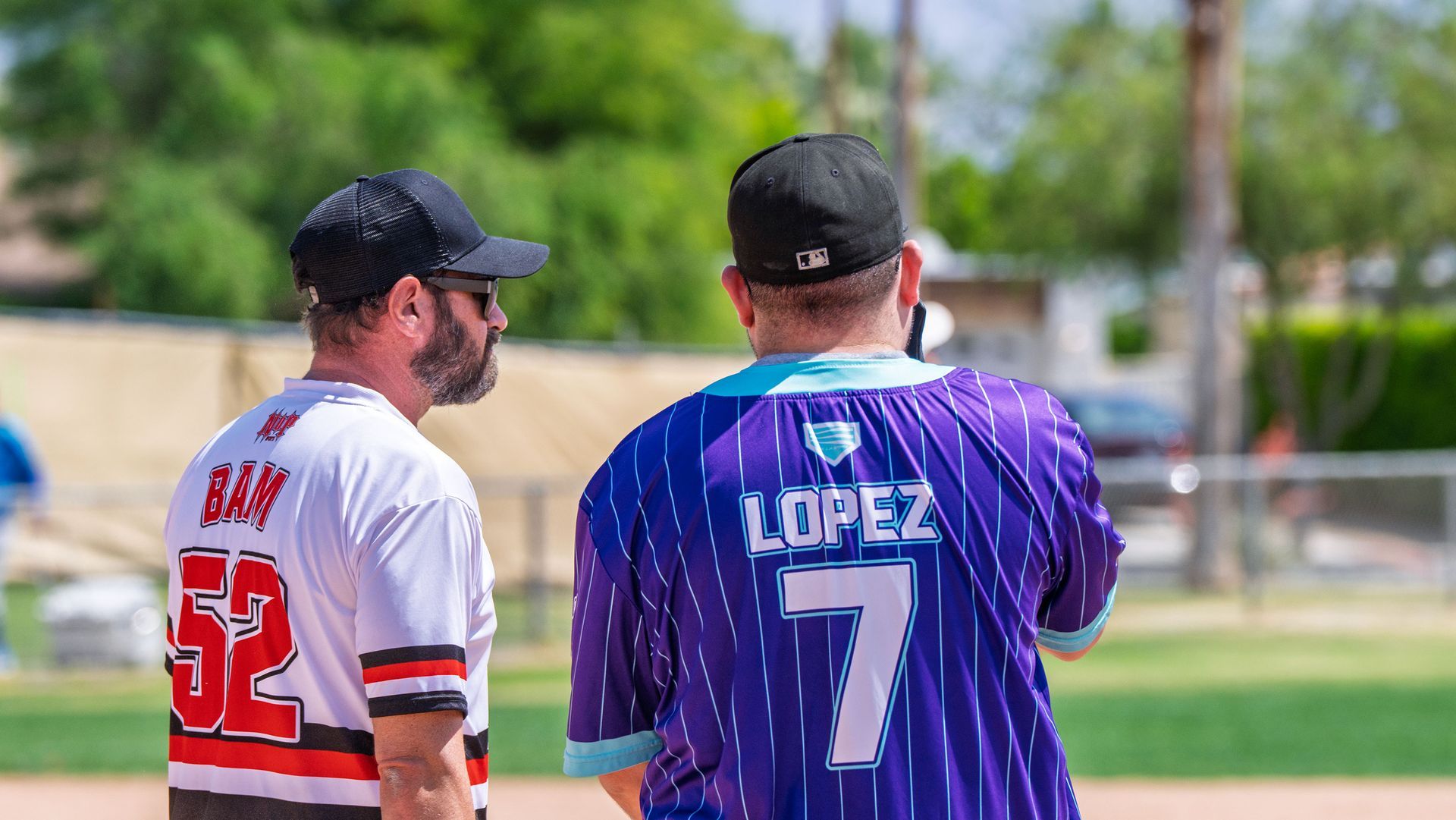 Two baseball players in jerseys, one in white #52 and one in purple #7, stand on a field talking.
