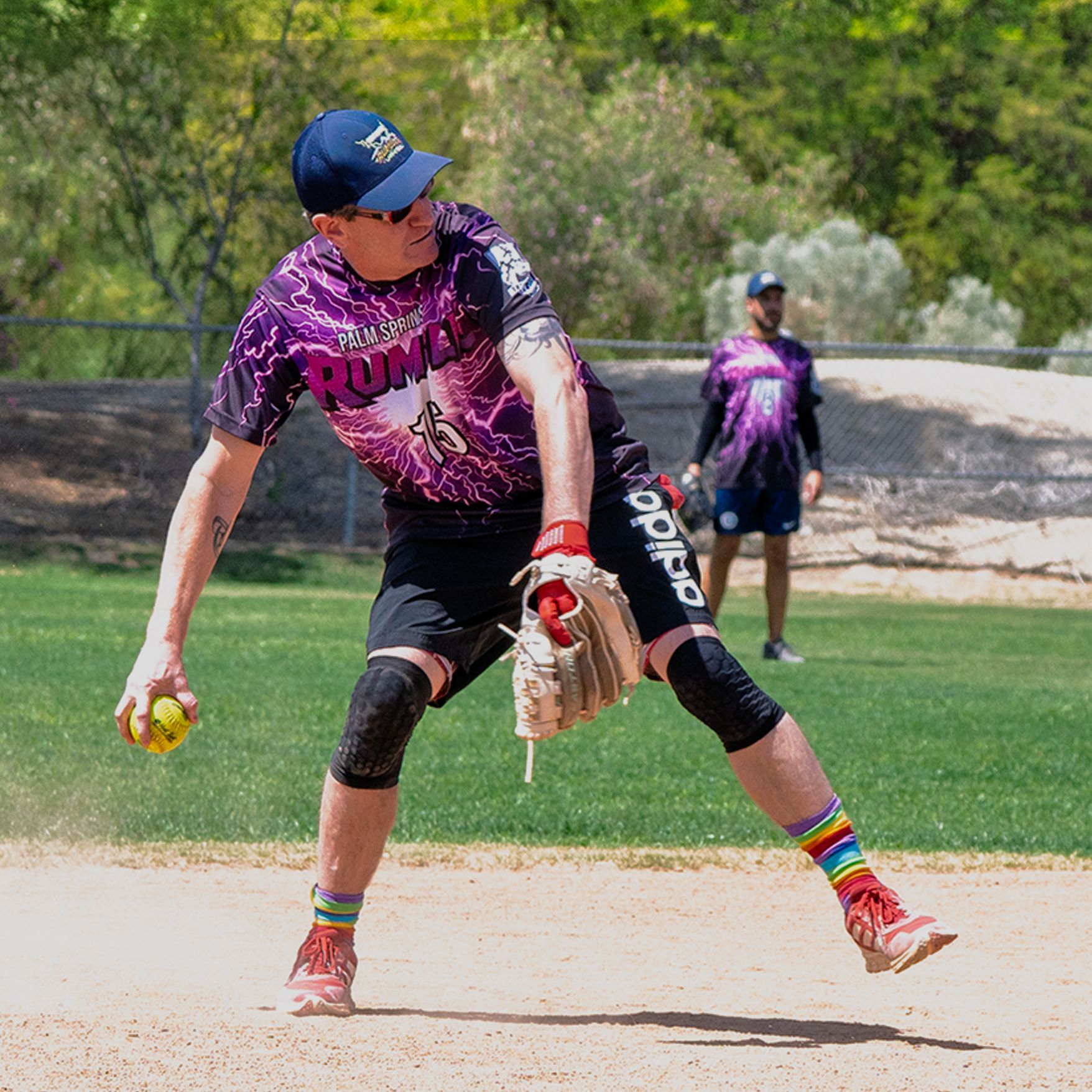 Softball player in purple uniform fielding a ball on a dirt infield, teammate in the background.