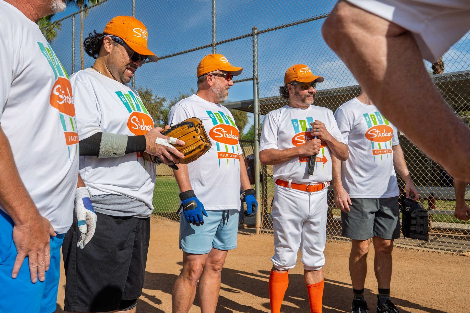 People in orange hard hats and gloves stand at a baseball field, holding a glove and watching attentively.
