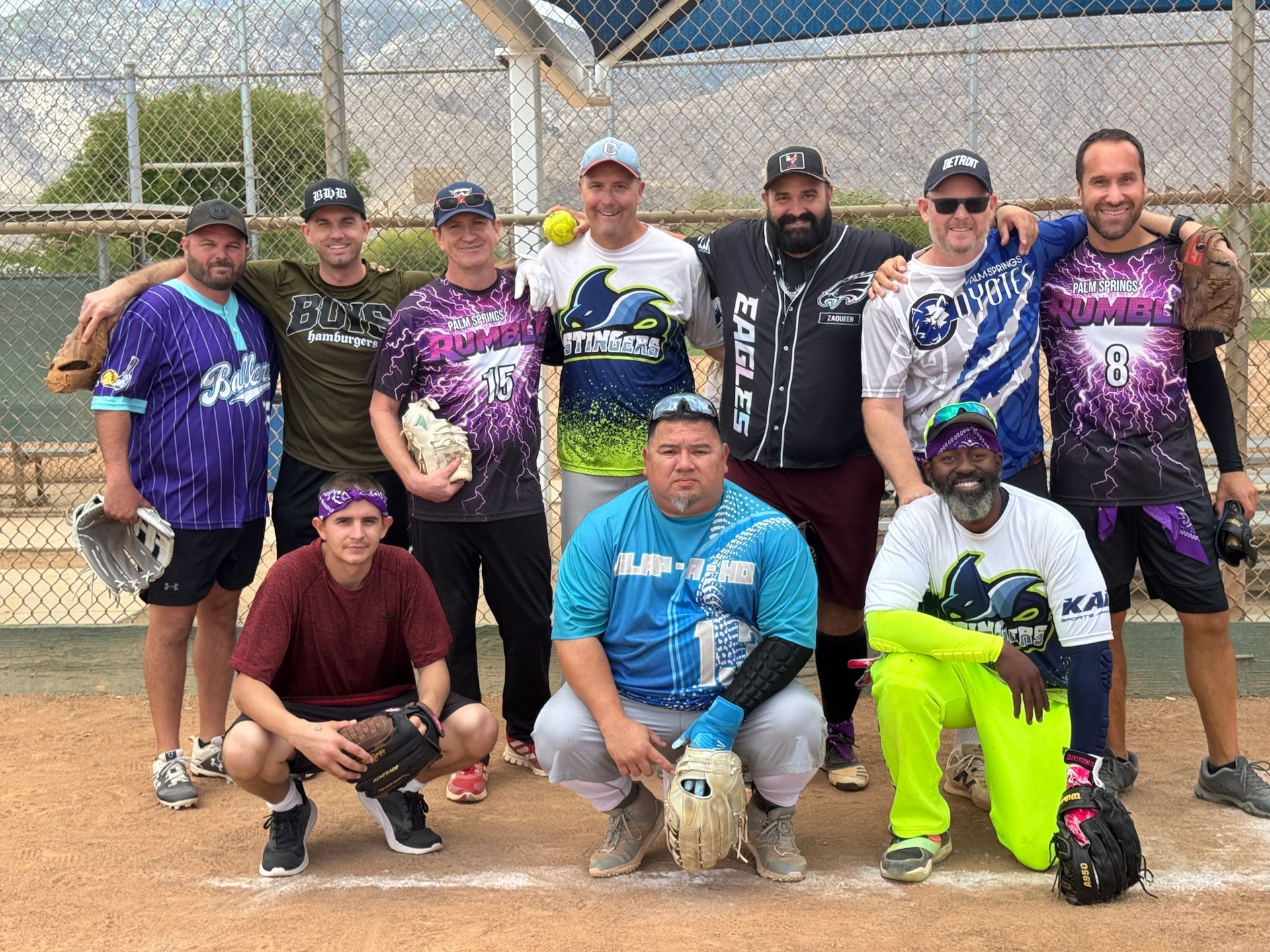 Group of 10 people posing in front of a chain-link fence on a dirt field, wearing colorful sports jerseys.