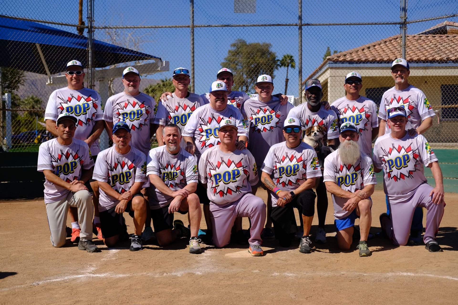 Youth baseball team posing for a group photo on a dirt field under a clear blue sky.