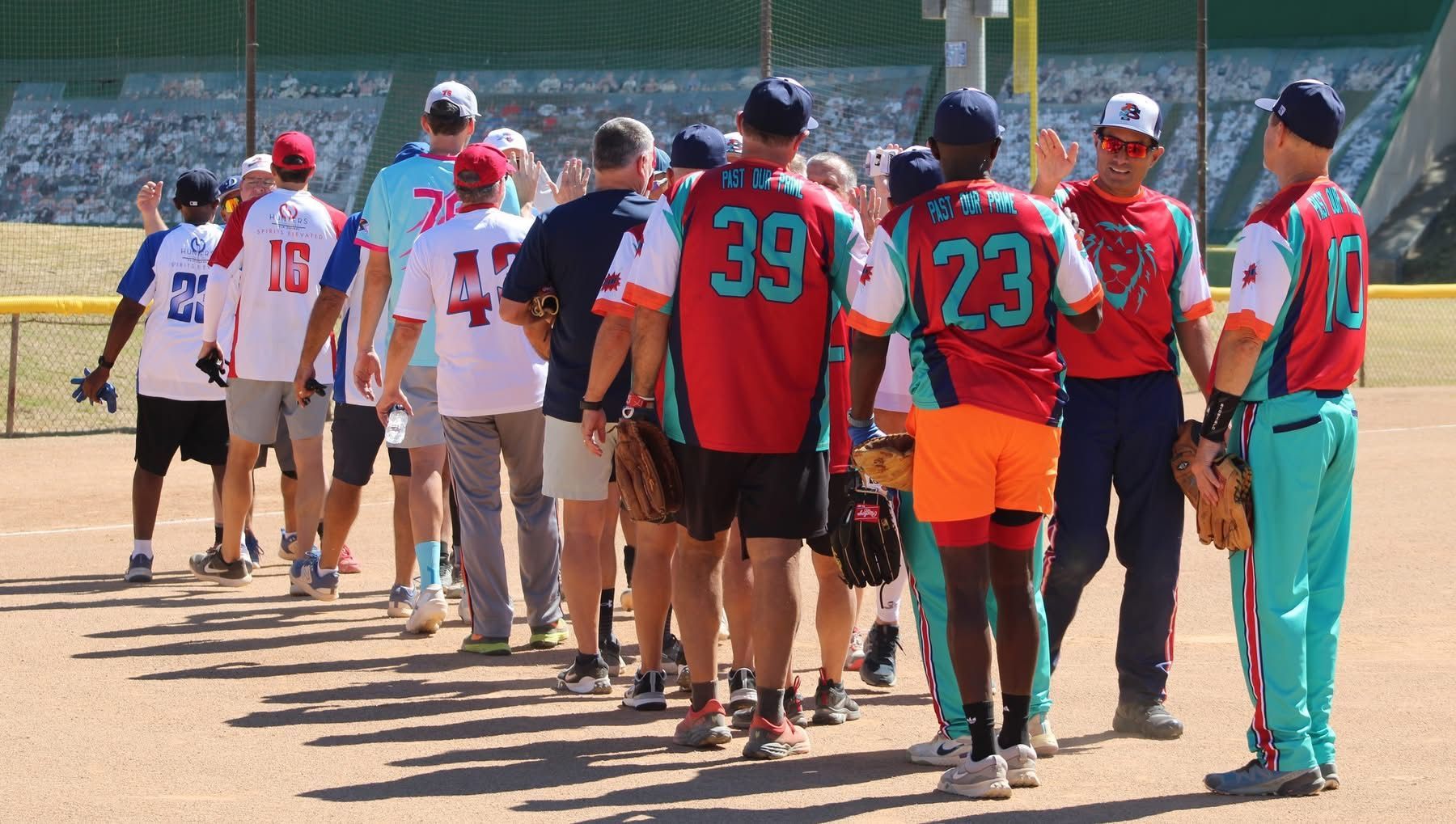 People in red and teal sports jerseys standing in a line on a dusty field during a game