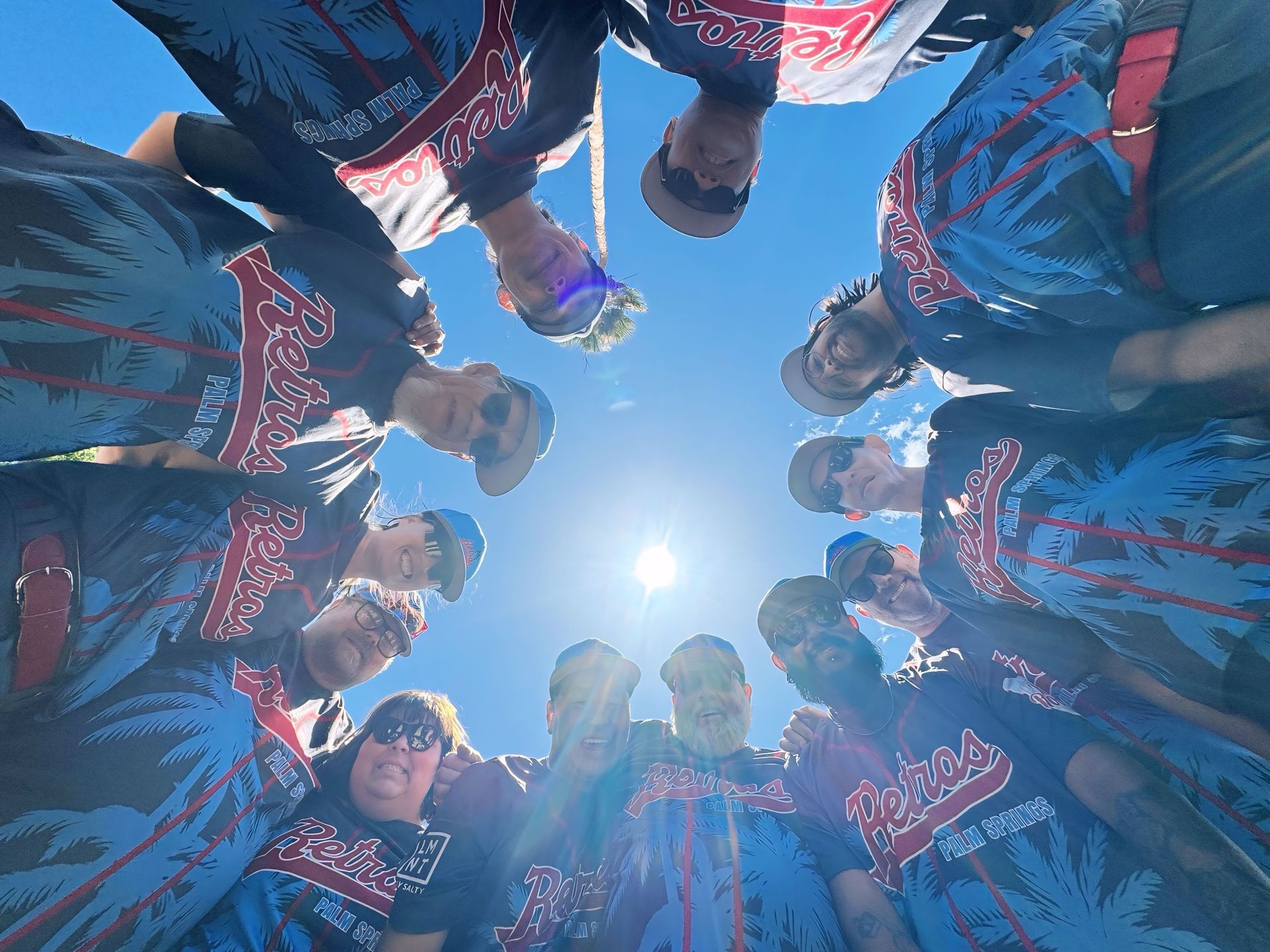People in blue-and-red jackets standing in a circle, looking down at the camera against a bright sky.