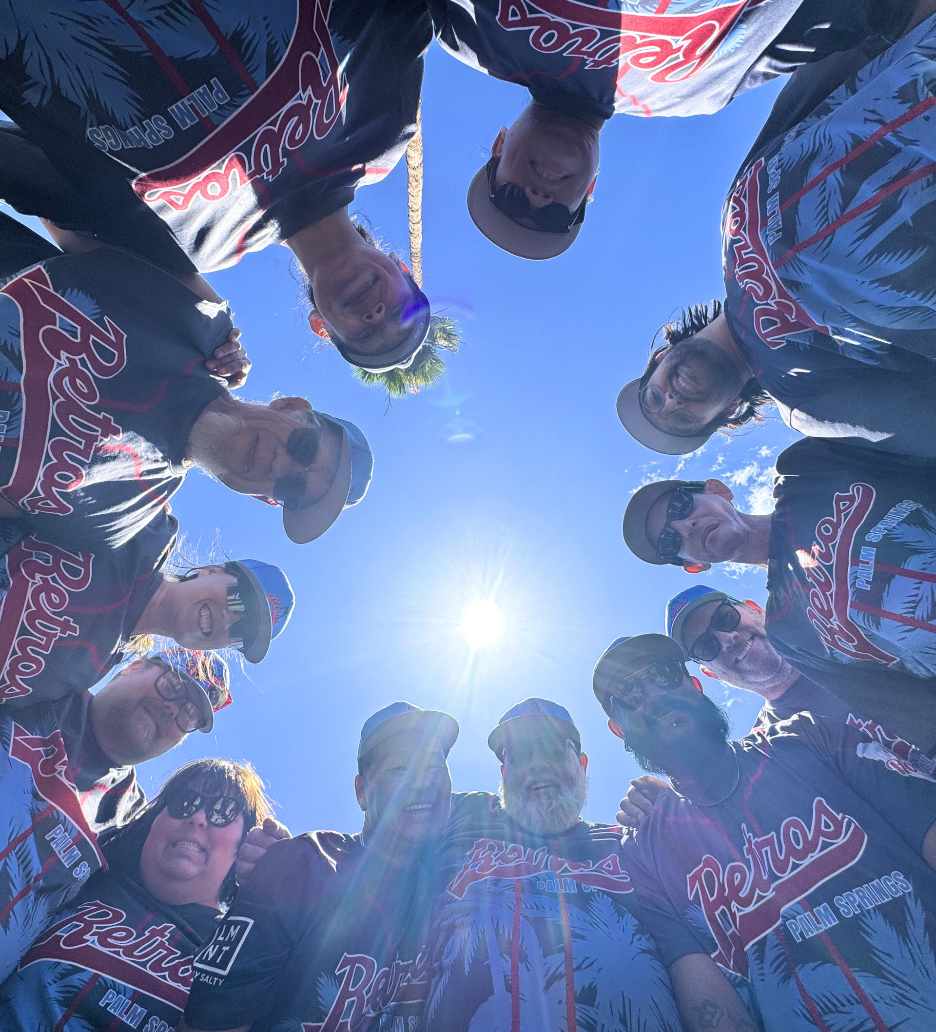 Cyclists in matching jerseys huddle in a circle, looking down toward the camera against a bright blue sky.