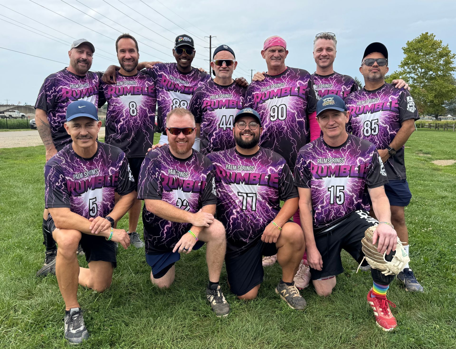 Group of runners in neon jerseys posing outdoors on grass, smiling and crouching together before a race.