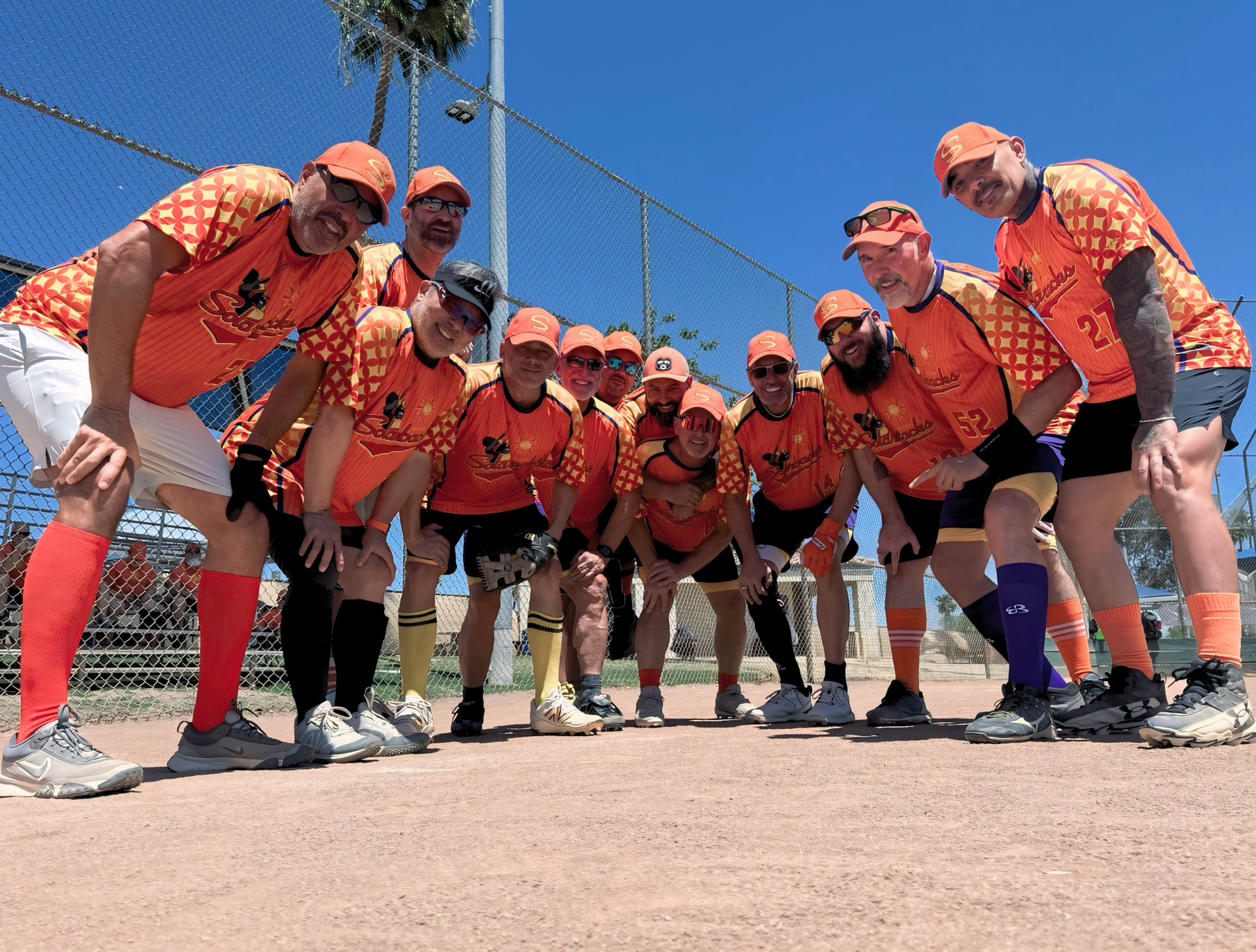 Softball team in orange jerseys posing together on a sunny field, leaning toward the camera.