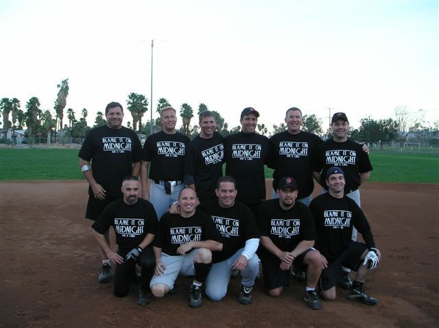 Group of people in matching black shirts posing on a baseball field at dusk.