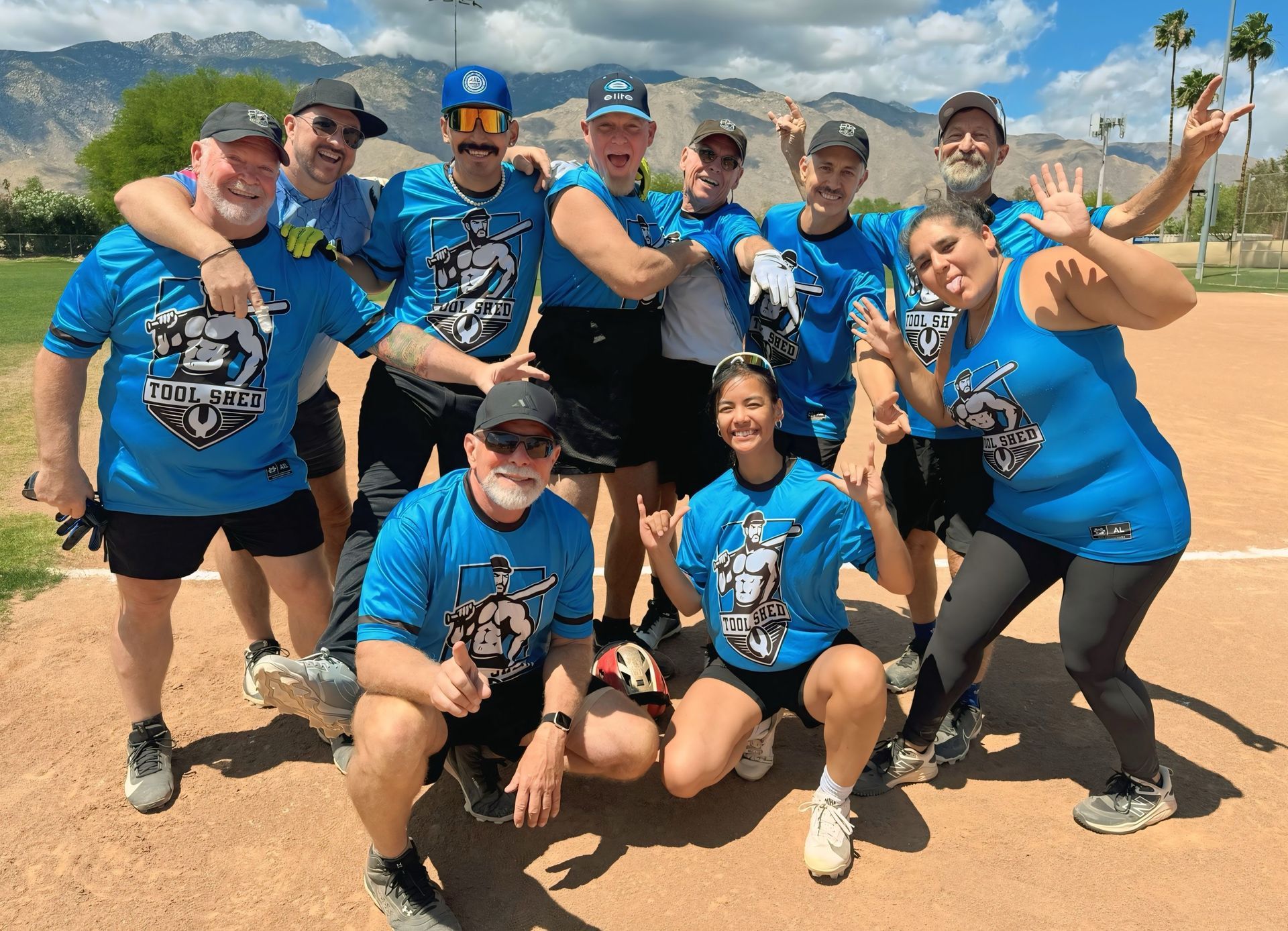 Group of teammates in blue shirts posing together on a baseball field, smiling and cheering.