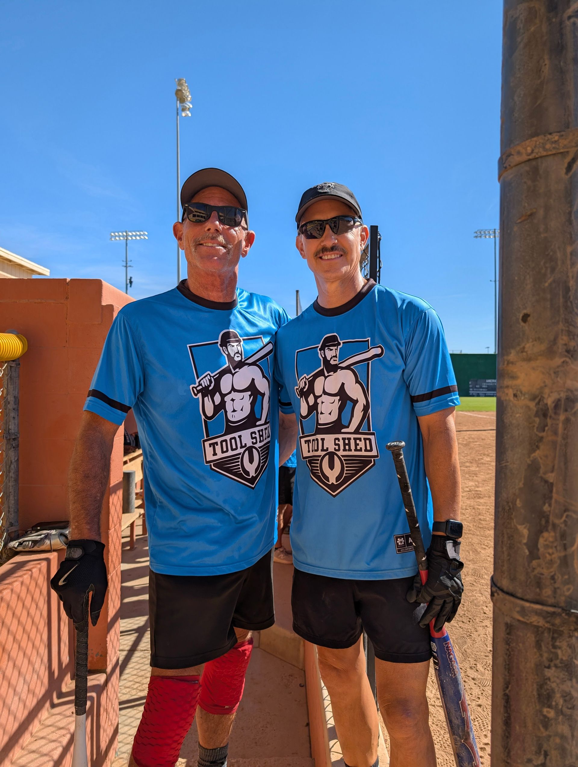 Two people in matching blue shirts and sunglasses pose outdoors under a clear sky.