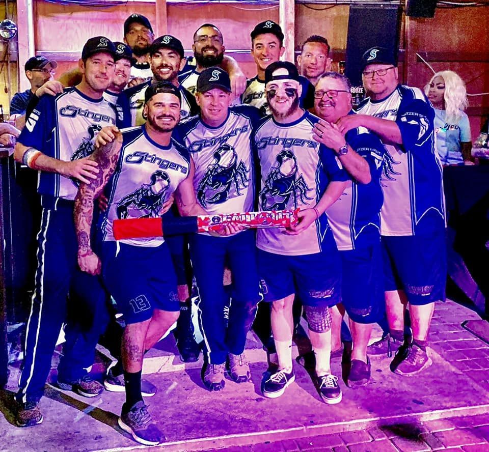 Group of cyclists posing together indoors, wearing matching jerseys and medals under purple lighting.