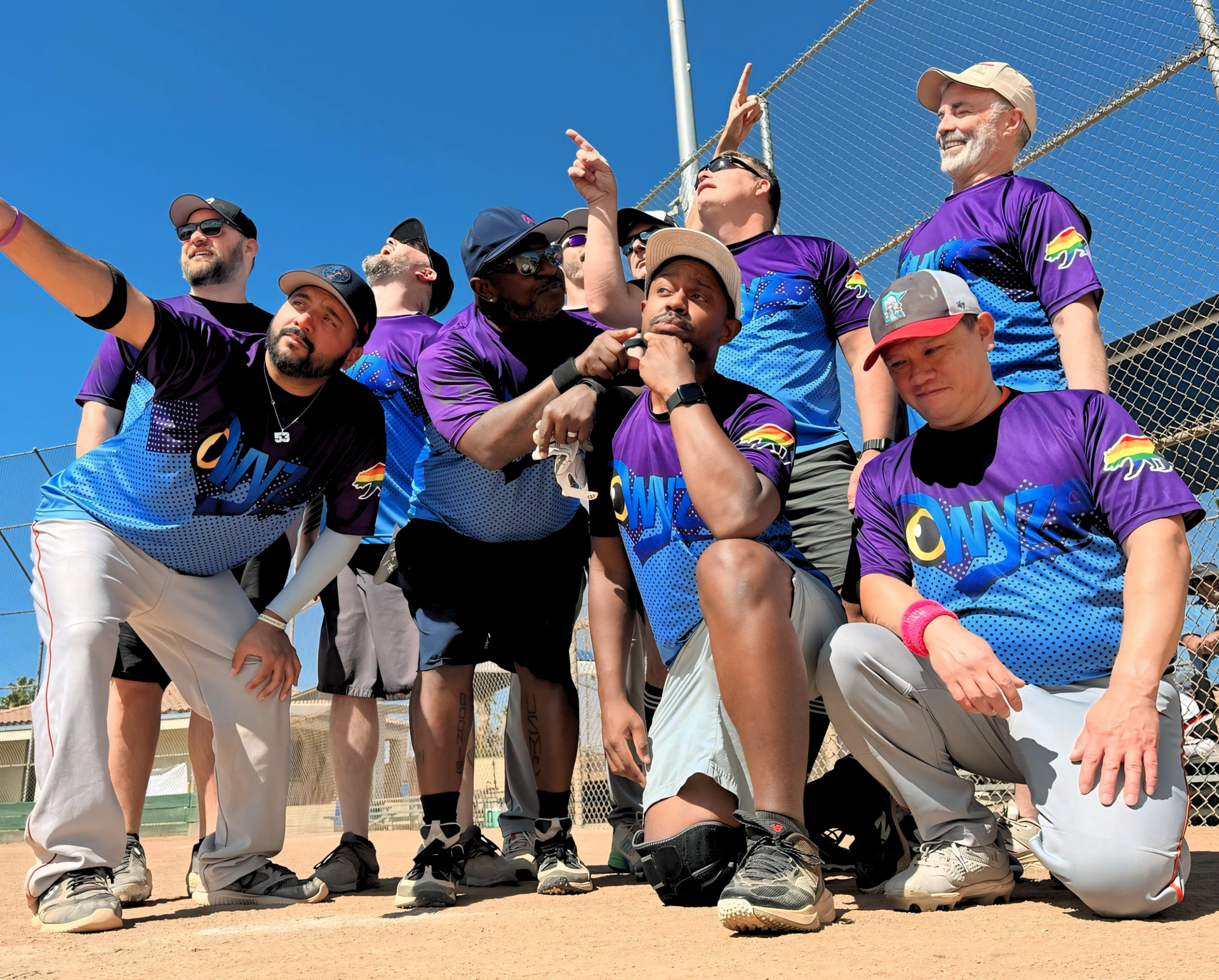 Group of people in matching purple and blue jerseys posing outdoors, pointing and kneeling on a sunny field.