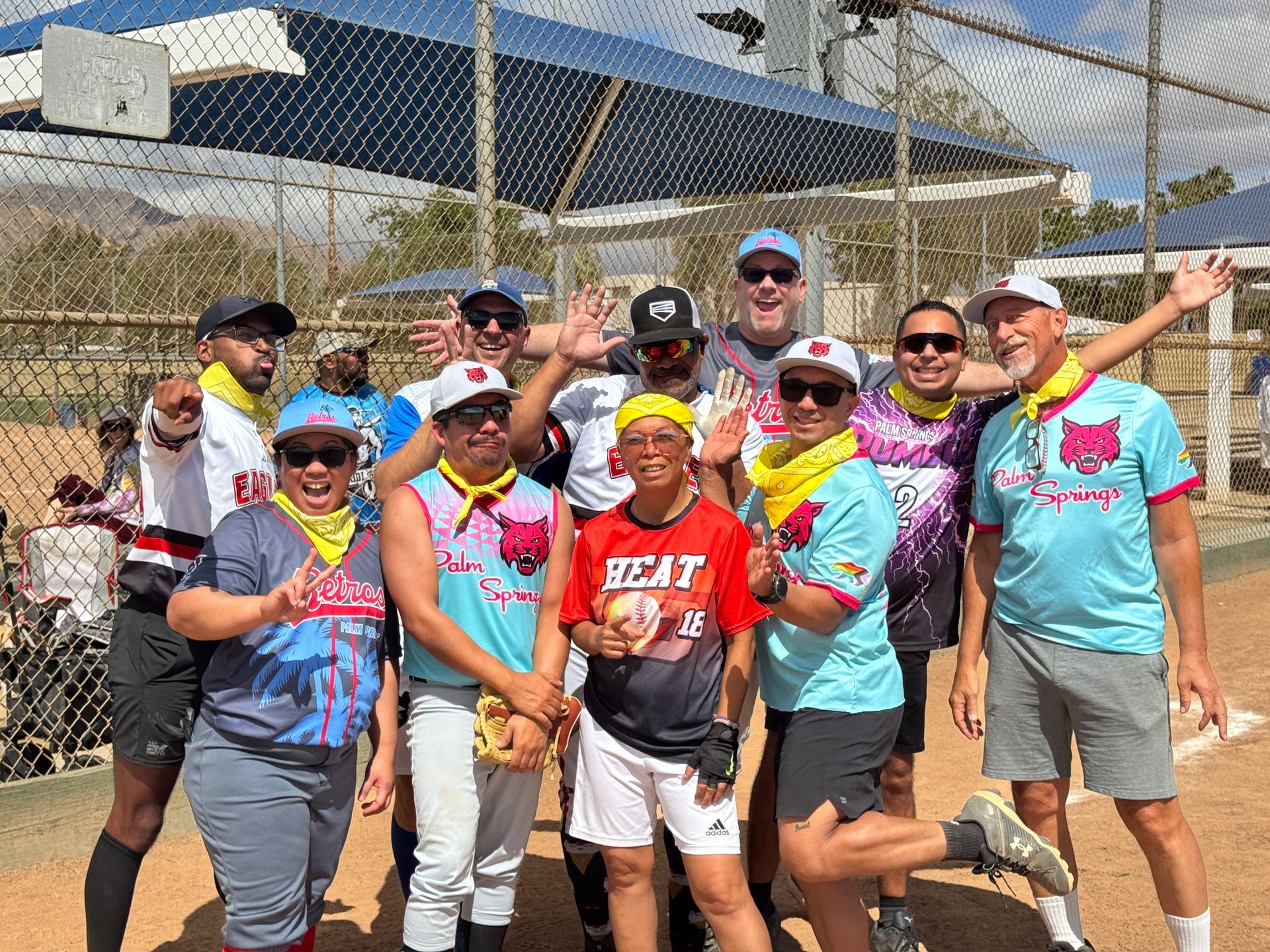 Group of smiling players in colorful jerseys posing on a softball field behind a chain-link fence.