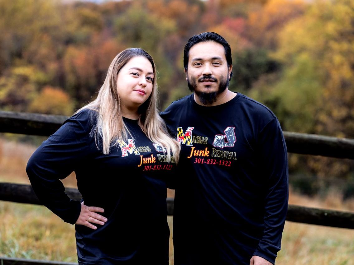 Couple wearing matching black shirts with logo, standing near a wooden fence outdoors, autumn foliage background.